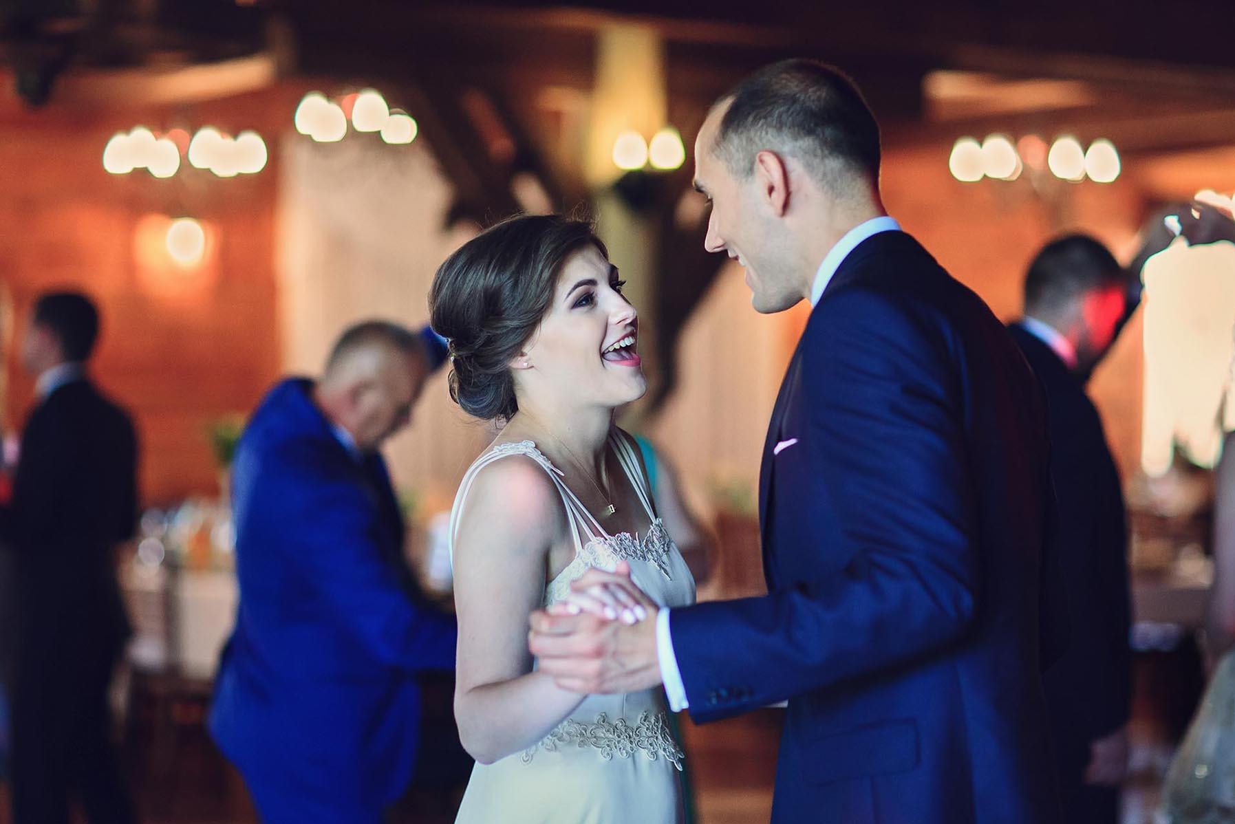 Uśmiechnięta panna młoda w kremowej sukni i pan młody w granatowym garniturze trzymają się za ręce podczas pierwszego tańca na sali weselnej z dekoracjami.