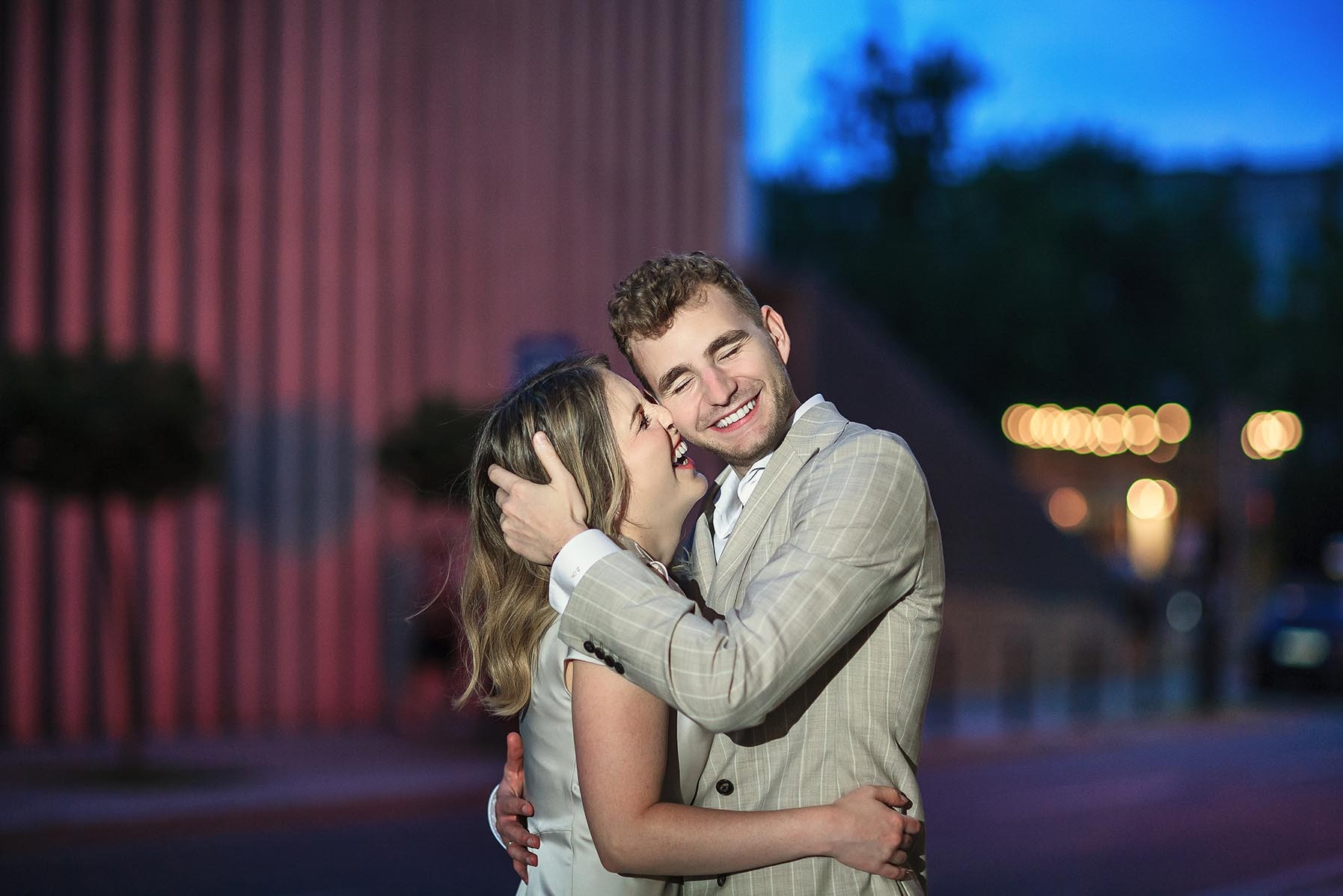 Para młoda w objęciach, śmiech na twarzach, wieczorne światło. Pan w garniturze w kratę, Panna Młoda w jasnej sukience. Romantyczna sesja w scenerii miejskiej.