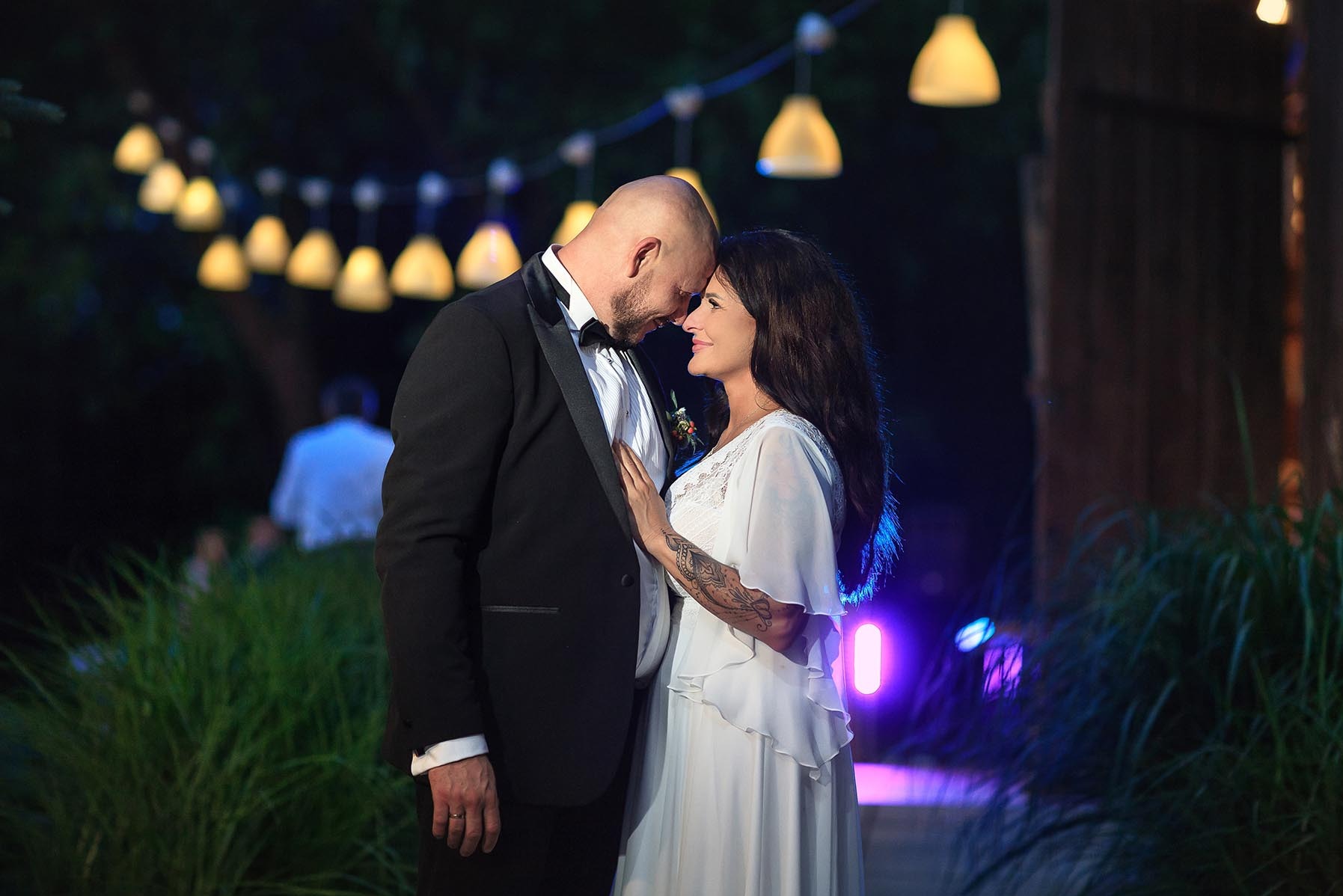 Para młoda, panna młoda w zwiewnej sukni z tatuażem na ramieniu, pan młody w smokingu, romantyczny kadr z oświetleniem girlandami, zbliżenie.