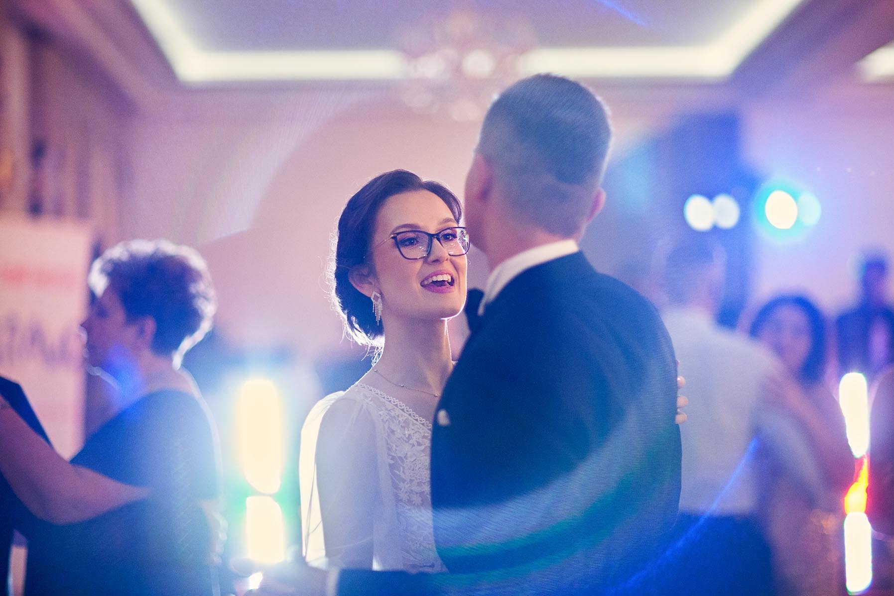 Uśmiechnięta panna młoda w okularach, wpatrzona w pana młodego podczas pierwszego tańca na sali weselnej, w tle goście i dekoracje.