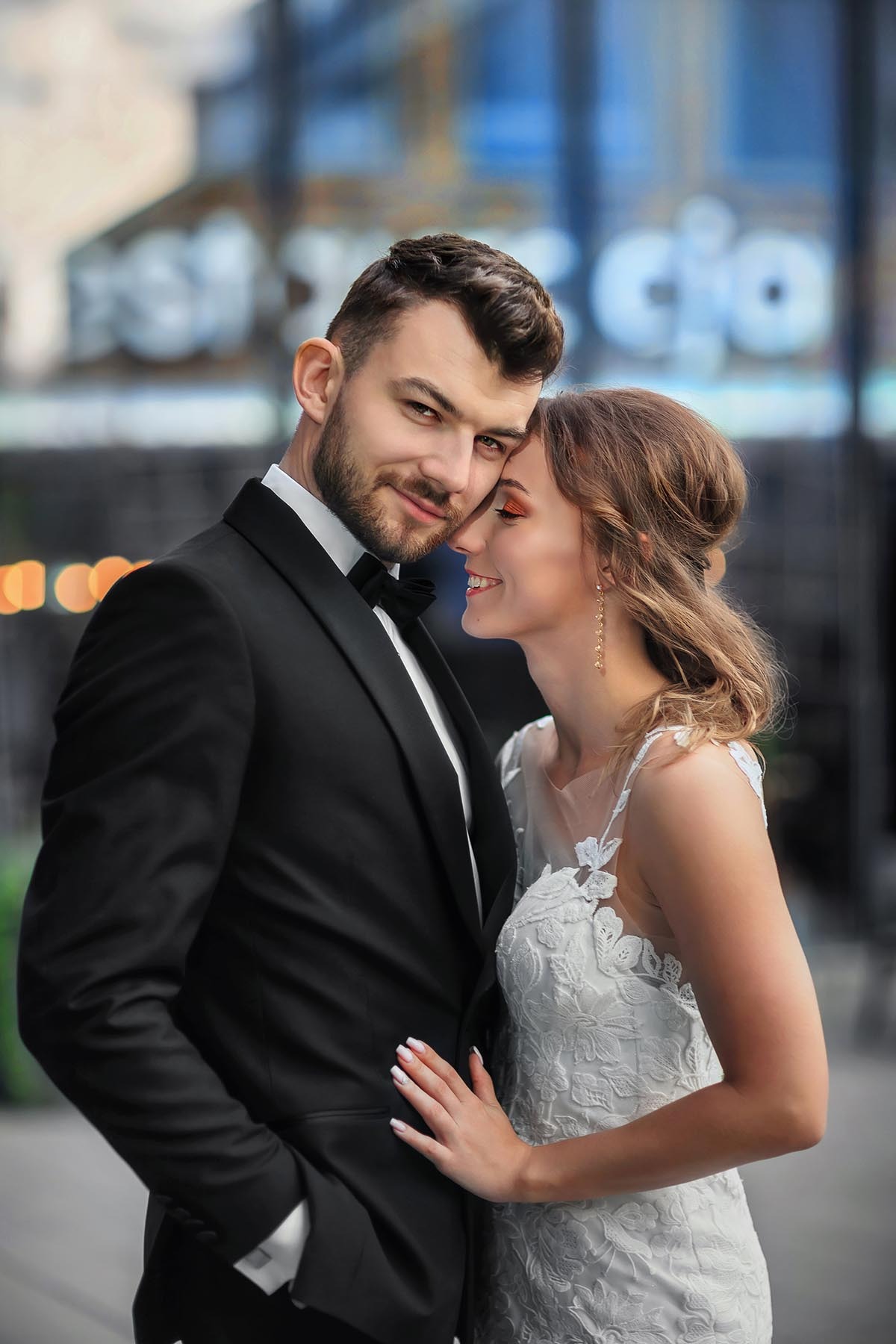 Uśmiechnięta para młoda: brunet w smokingu i panna młoda w koronkowej sukni, przytuleni na tle miejskich świateł. Romantyczny portret ślubny.
