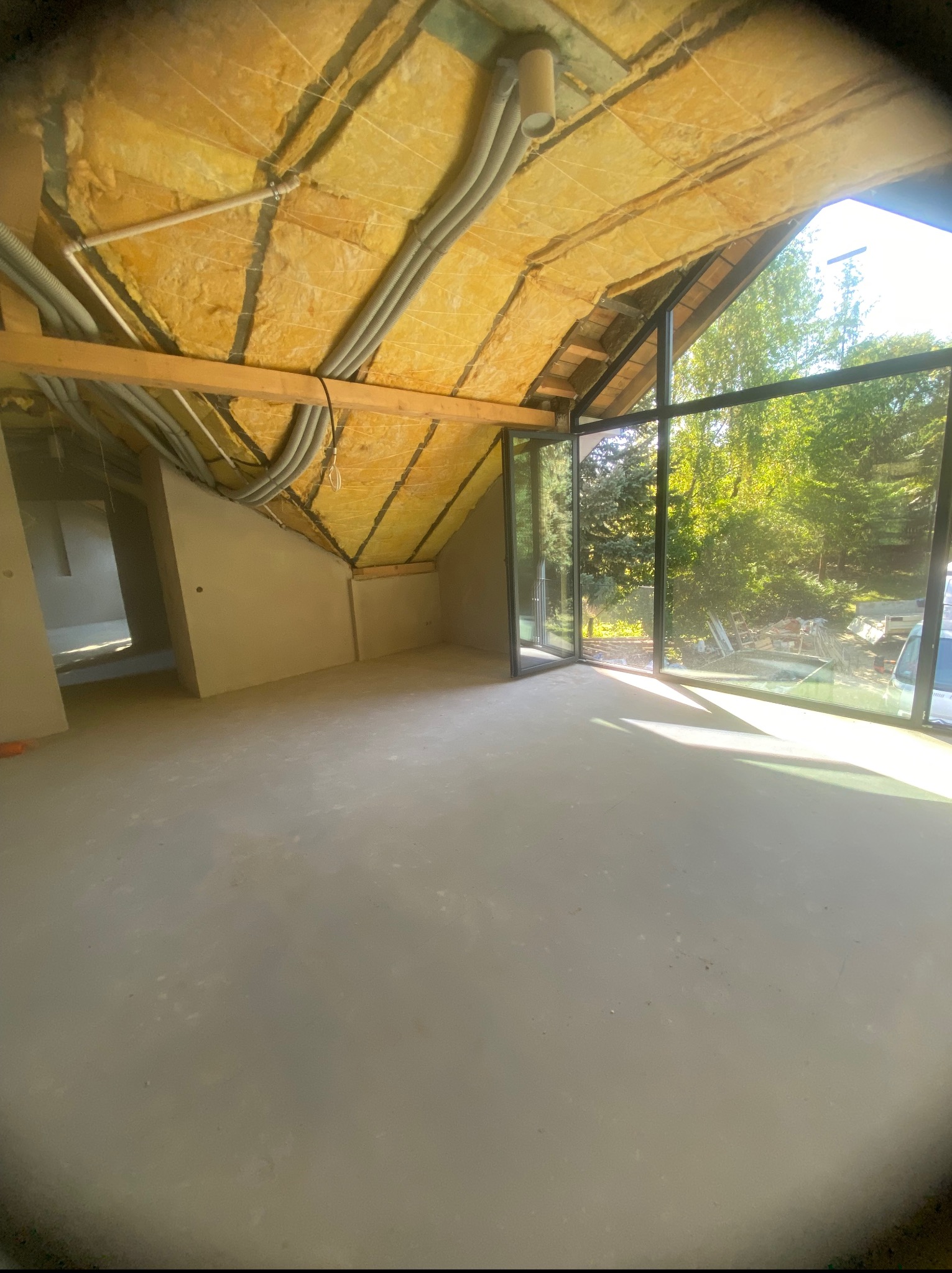 Surowe wnętrze poddasza z oknami panoramicznymi i ociepleniem stropu wełną mineralną. Widoczne instalacje elektryczne i ściany przygotowane pod wykończenie.