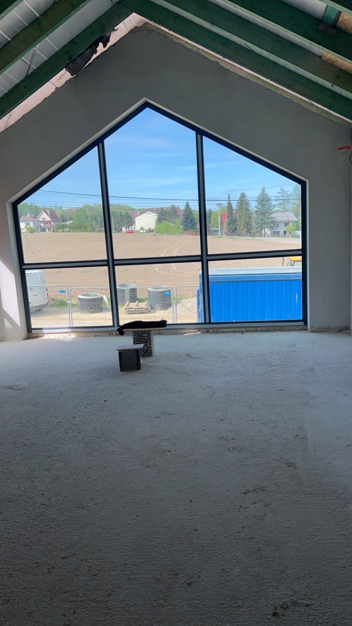 Surowe wnętrze poddasza z dużym oknem w kształcie trapezu, widok na pole i zabudowania. Ściany przygotowane do wykończenia, surowa podłoga, widoczne elementy konstrukcji dachu.