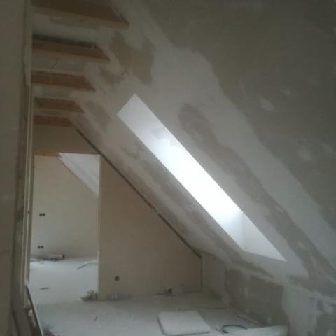 Poddasze po szpachlowaniu, widoczne okno dachowe i fragment drewnianej konstrukcji. Ściany przygotowane do malowania, widoczne ślady po szpachli.