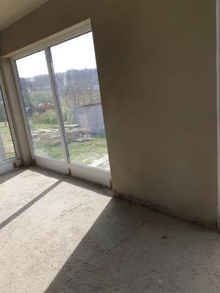 Wnętrze w stanie surowym: ściana po tynkowaniu, duże okno z widokiem na zewnątrz, betonowa podłoga z cieniem. Widoczne niedoskonałości i surowość wykończenia.