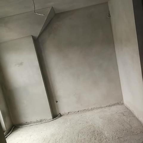 Wnętrze po tynkowaniu maszynowym: gładkie, szare ściany i sufit, widoczne przewody elektryczne przy podłodze. Minimalistyczne wykończenie, surowy stan.