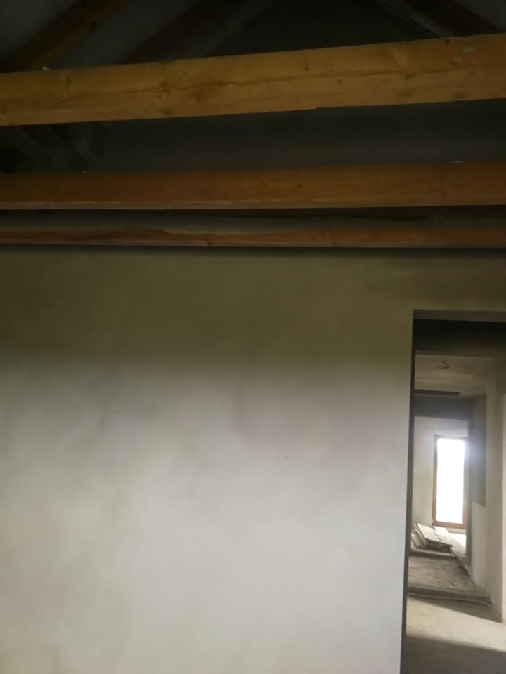 Świeżo otynkowana ściana wewnętrzna z widocznymi drewnianymi belkami stropowymi i prześwitem do kolejnego pomieszczenia z oknem w tle. Jasne, naturalne światło.