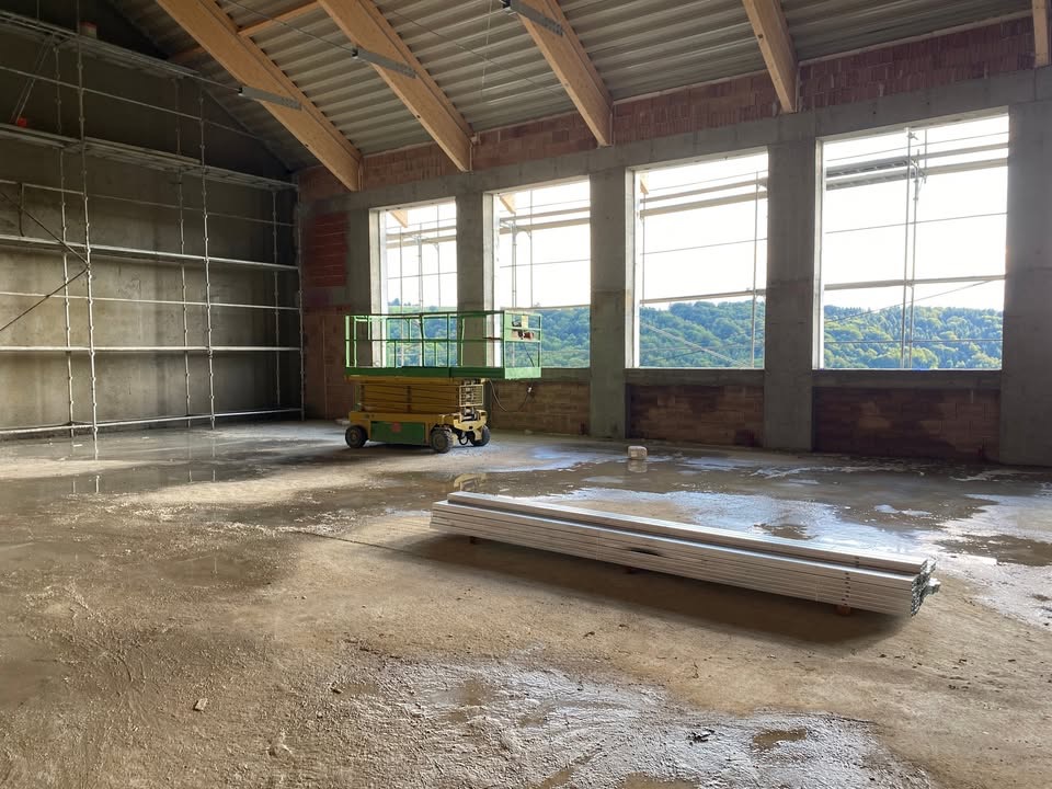 Wnętrze w trakcie prac tynkarskich: rusztowania, podnośnik nożycowy, surowe ściany z oknami i widokiem na zieleń. Na podłodze woda i profile.