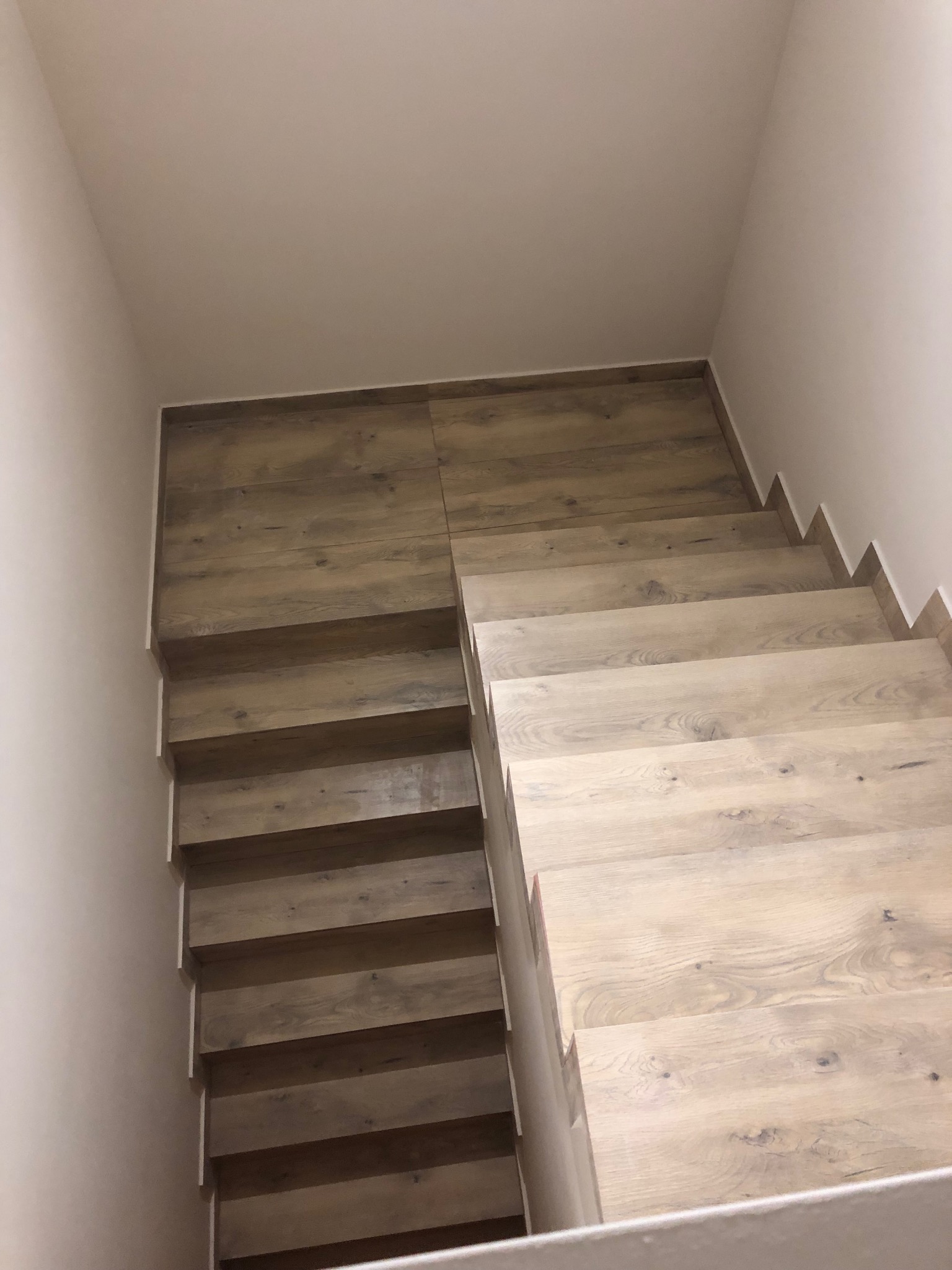 Drewniane schody w jasnym odcieniu, widok z góry, z wyraźnym rysunkiem słojów drewna. Minimalistyczne wykończenie ścian w neutralnym kolorze.