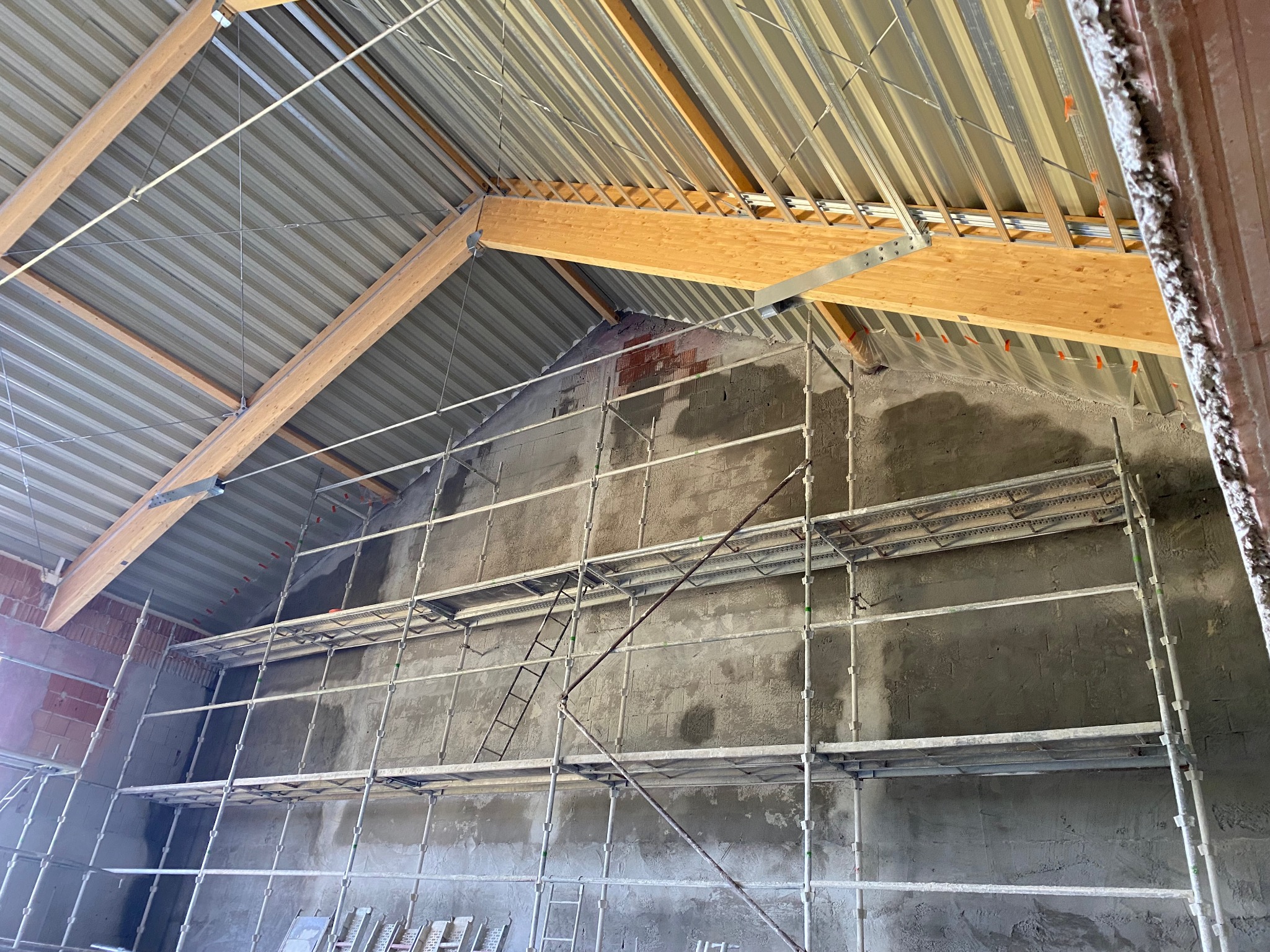 Wnętrze w trakcie remontu: surowa ściana z nałożonym tynkiem, rusztowanie, drewniana konstrukcja dachu z metalowym pokryciem. Widok z dołu, oświetlenie naturalne.
