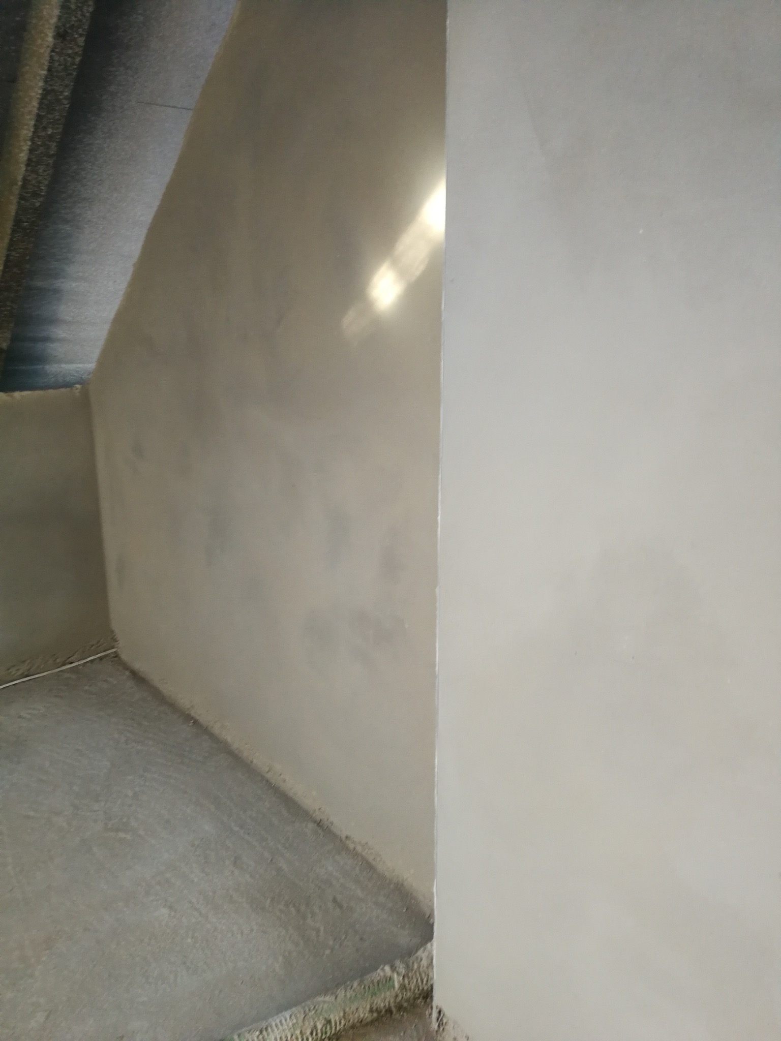 Świeżo położona, gładka, szara gładź na ścianach w rogu pomieszczenia. Widoczne refleksy światła na powierzchni. Podłoga również pokryta szarą warstwą.