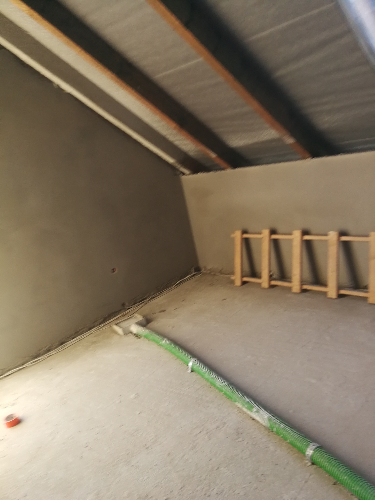 Świeżo otynkowane poddasze ze skośnym sufitem, widoczny przewód wentylacyjny i drewniana konstrukcja. Ściany gładkie, szare, gotowe do dalszych prac wykończeniowych.