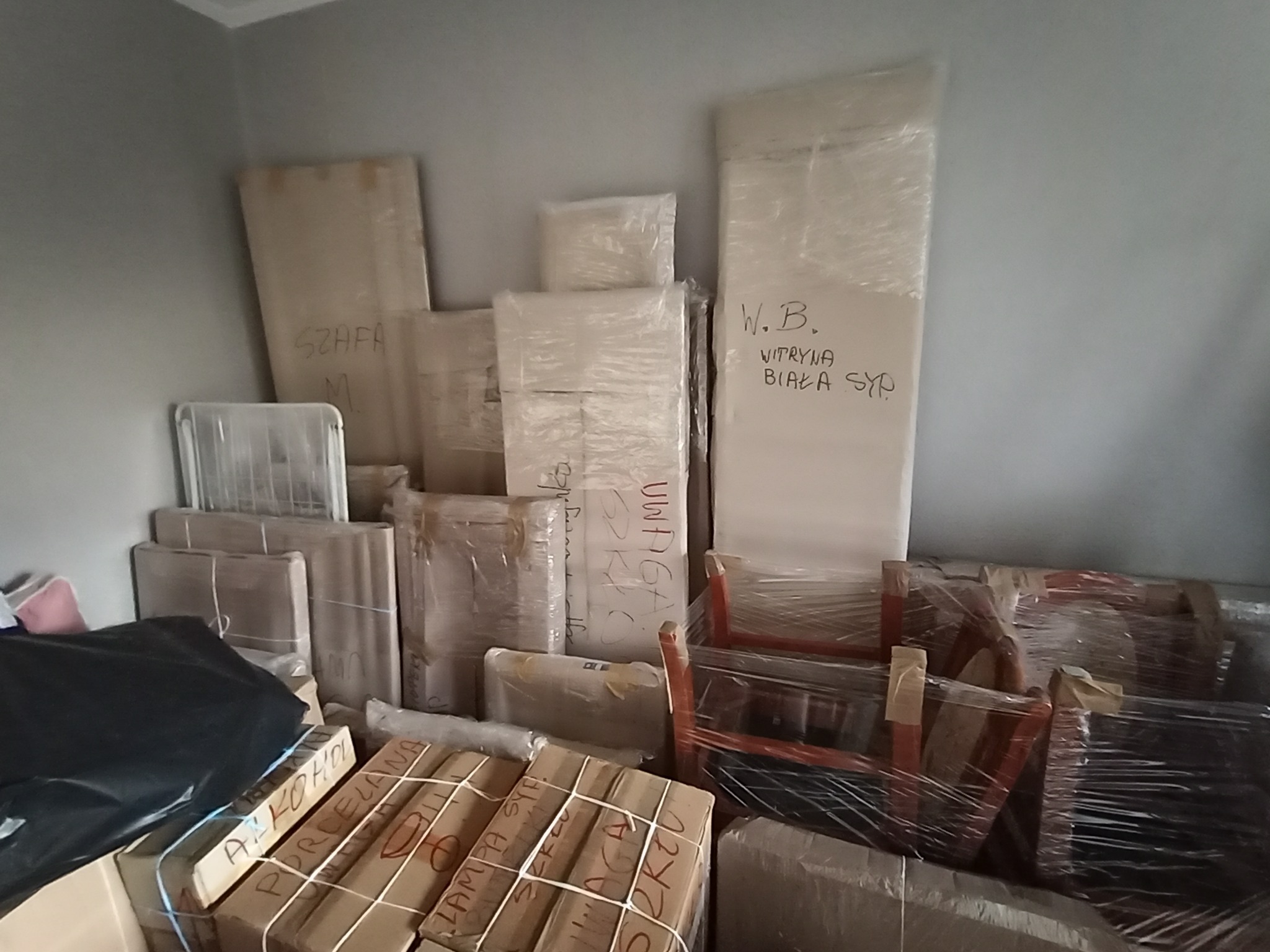 Spakowane meble i przedmioty, gotowe do transportu. Na kartonach widoczne napisy 'Porcelana' i 'Szkło'. Meble owinięte folią stretch.