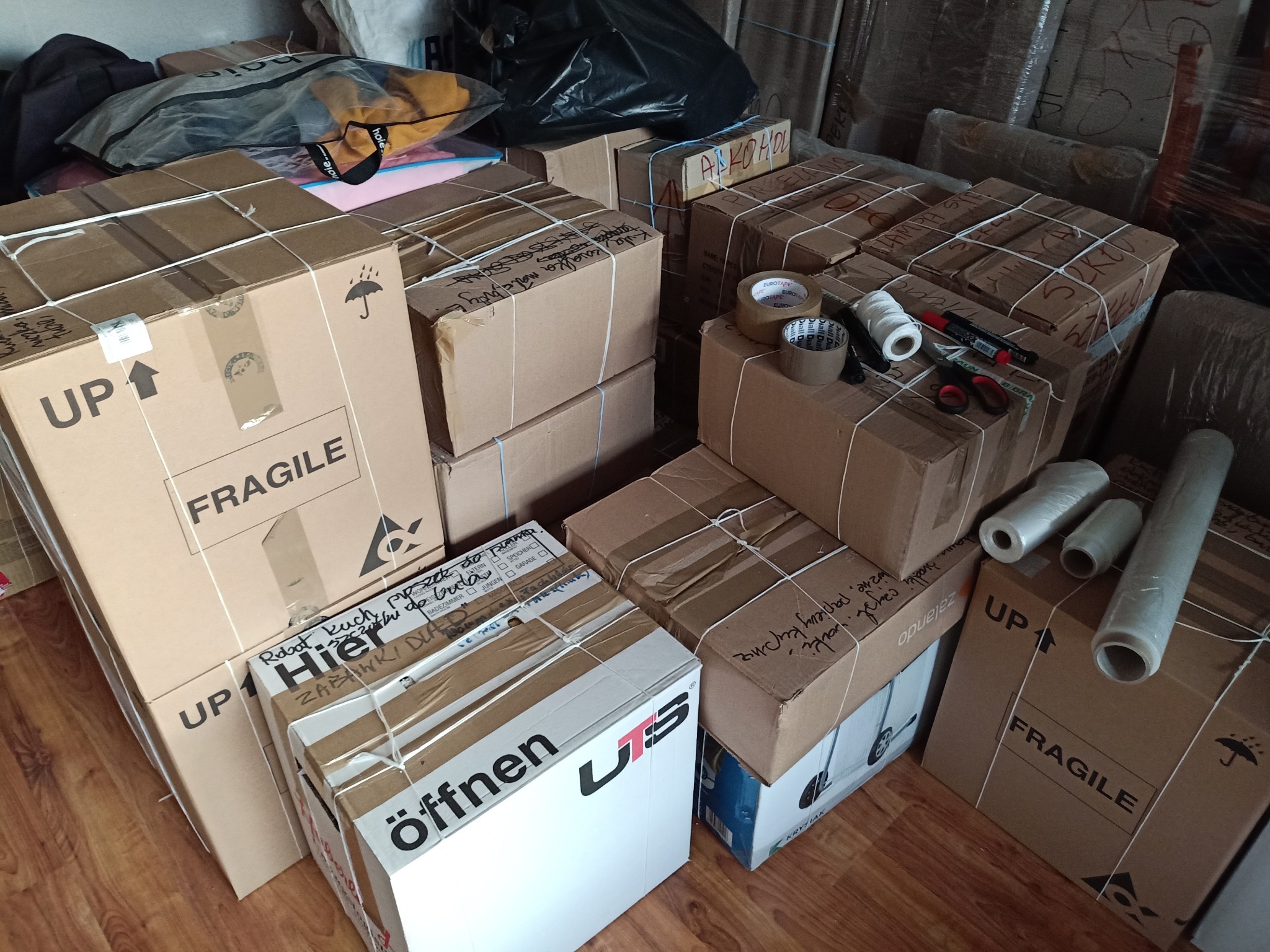 Spakowane i zabezpieczone kartony, gotowe do transportu. Widoczne akcesoria do pakowania: taśmy, sznurki, folia stretch i nożyczki. Na kartonach napisy ostrzegawcze i adresowe.