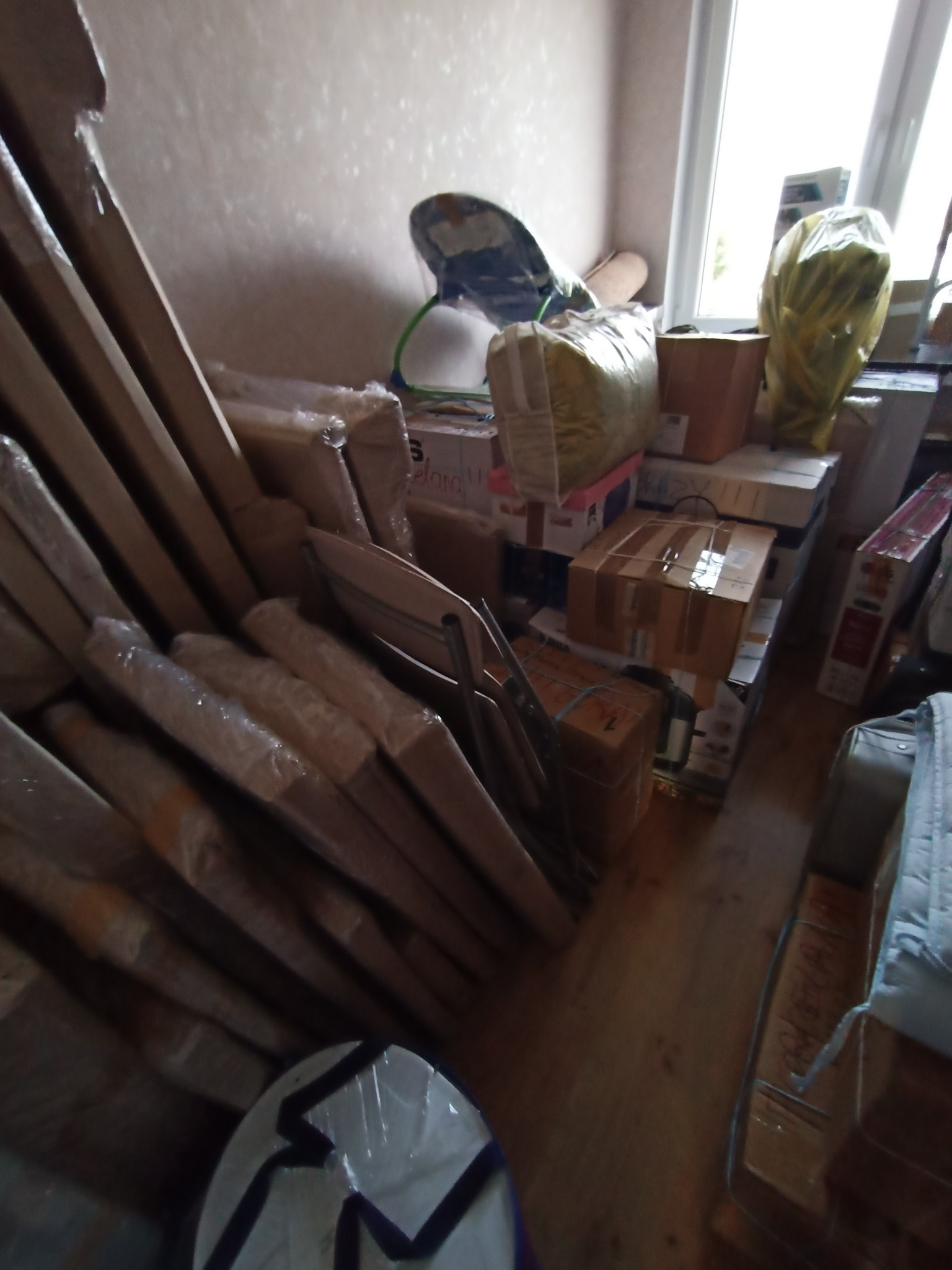 Spakowane kartony, meble i przedmioty w folii, gotowe do transportu. Widoczne okno w tle. Przygotowania do przeprowadzki.