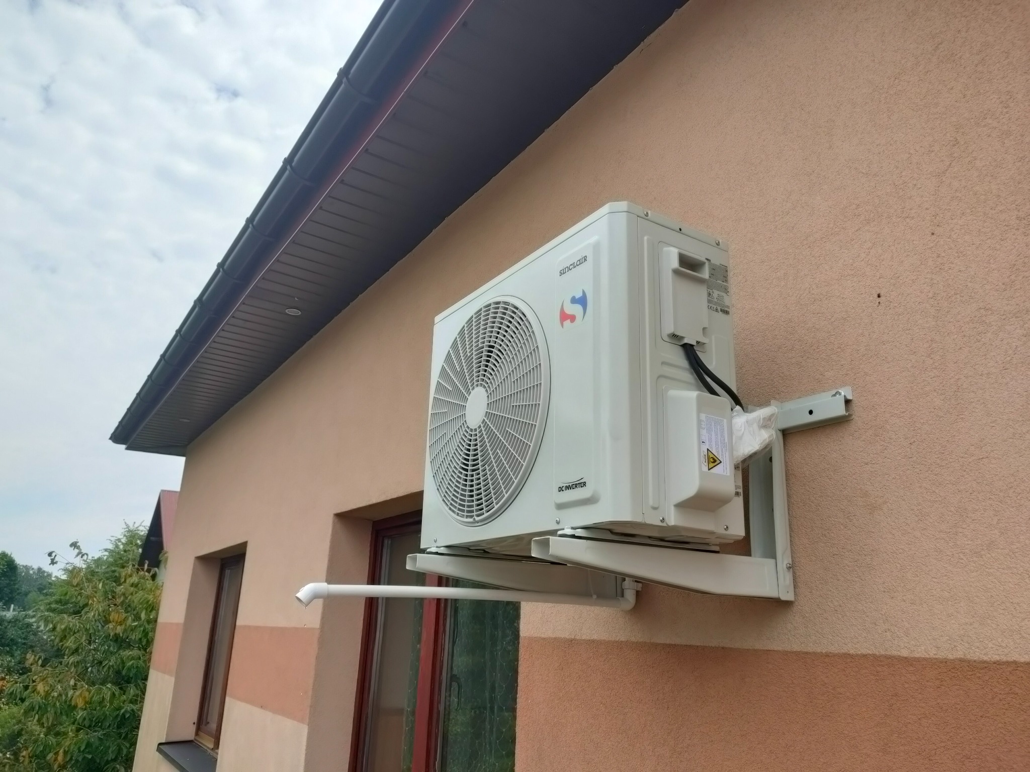 Biała jednostka zewnętrzna klimatyzacji marki Sinclair zamontowana na elewacji budynku z widoczną rurą odprowadzającą skropliny. Kątowe ujęcie z dołu.