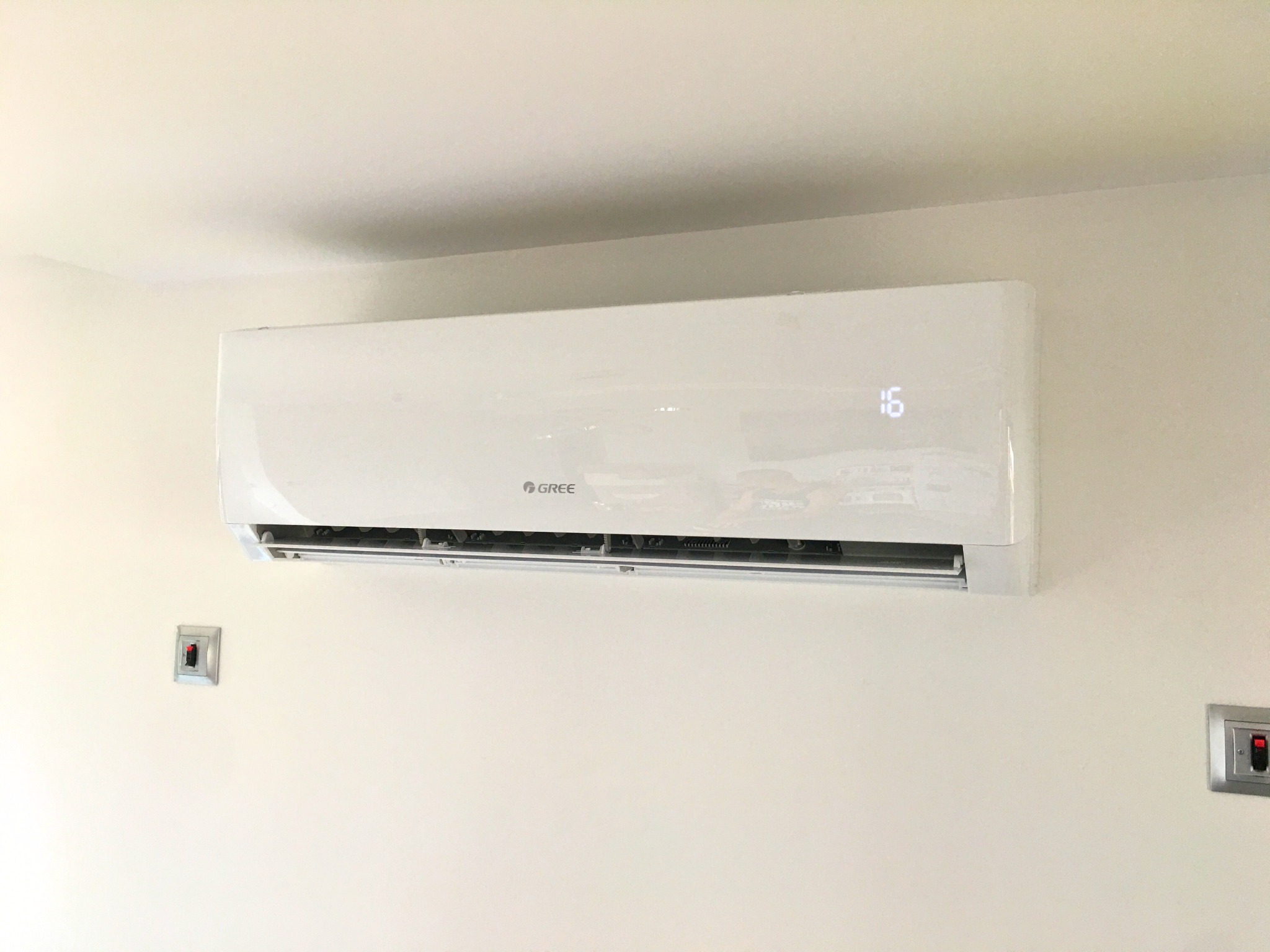 Biała klimatyzacja GREE zamontowana na jasnej ścianie, widoczne dwa włączniki. Na wyświetlaczu klimatyzacji widoczna temperatura 16 stopni.