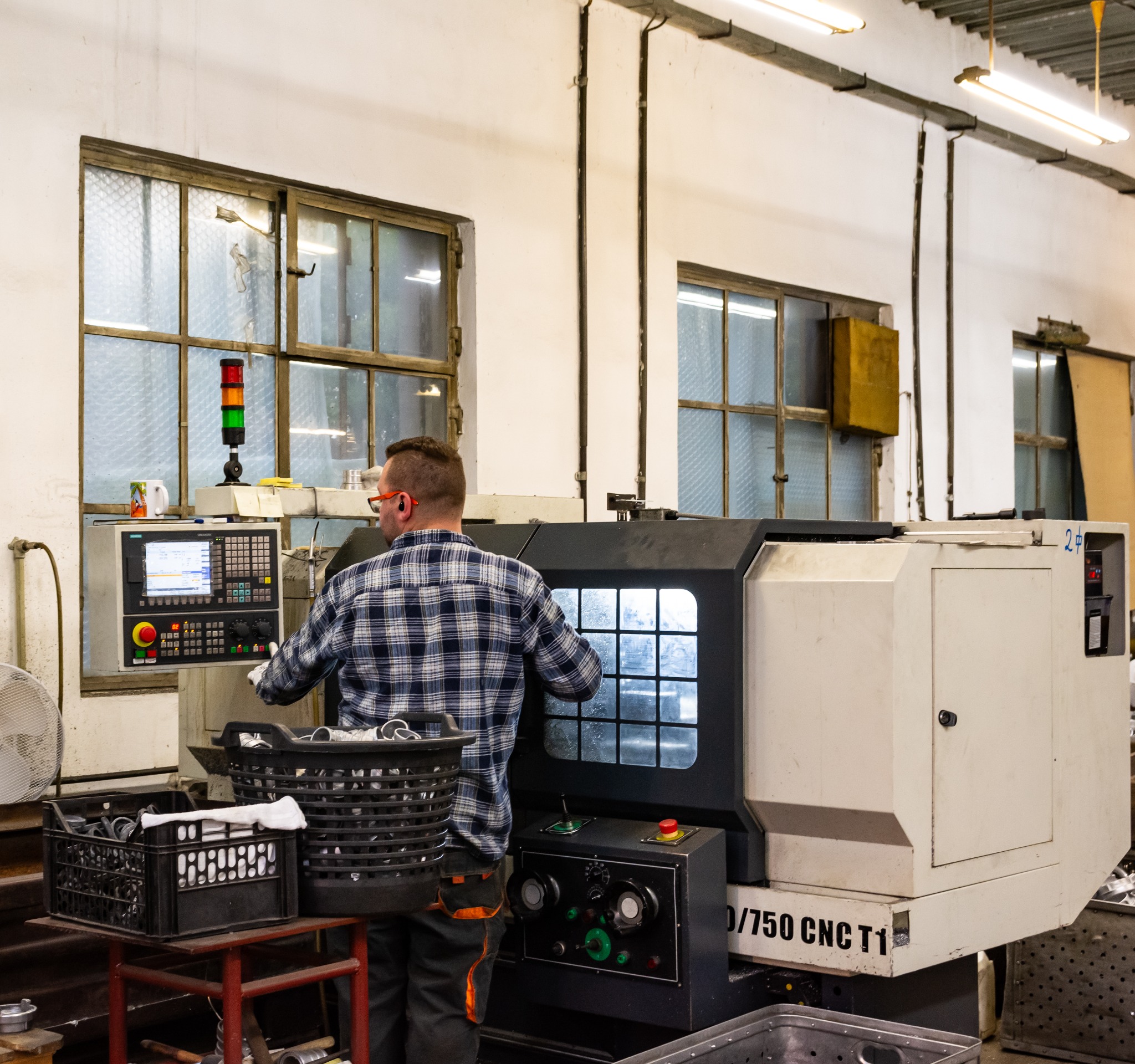 Pracownik w okularach ochronnych obsługuje tokarkę CNC w warsztacie. Widoczne okno i kosze z detalami. Oświetlenie jarzeniowe.