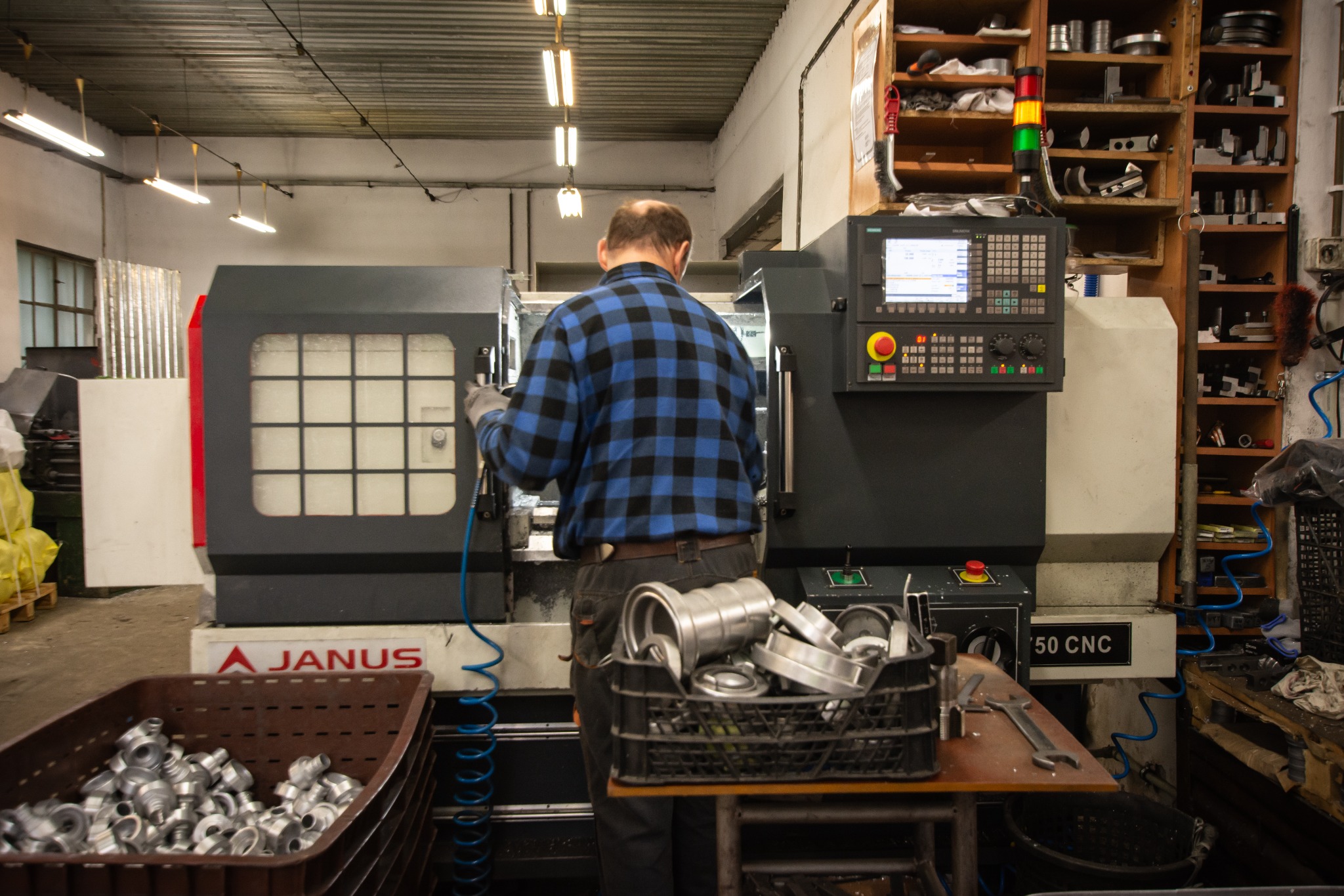 Pracownik w niebieskiej kracie obsługuje tokarkę CNC Janus. Metalowe elementy w koszach i na stole, oświetlenie przemysłowe. Warsztatowy klimat.