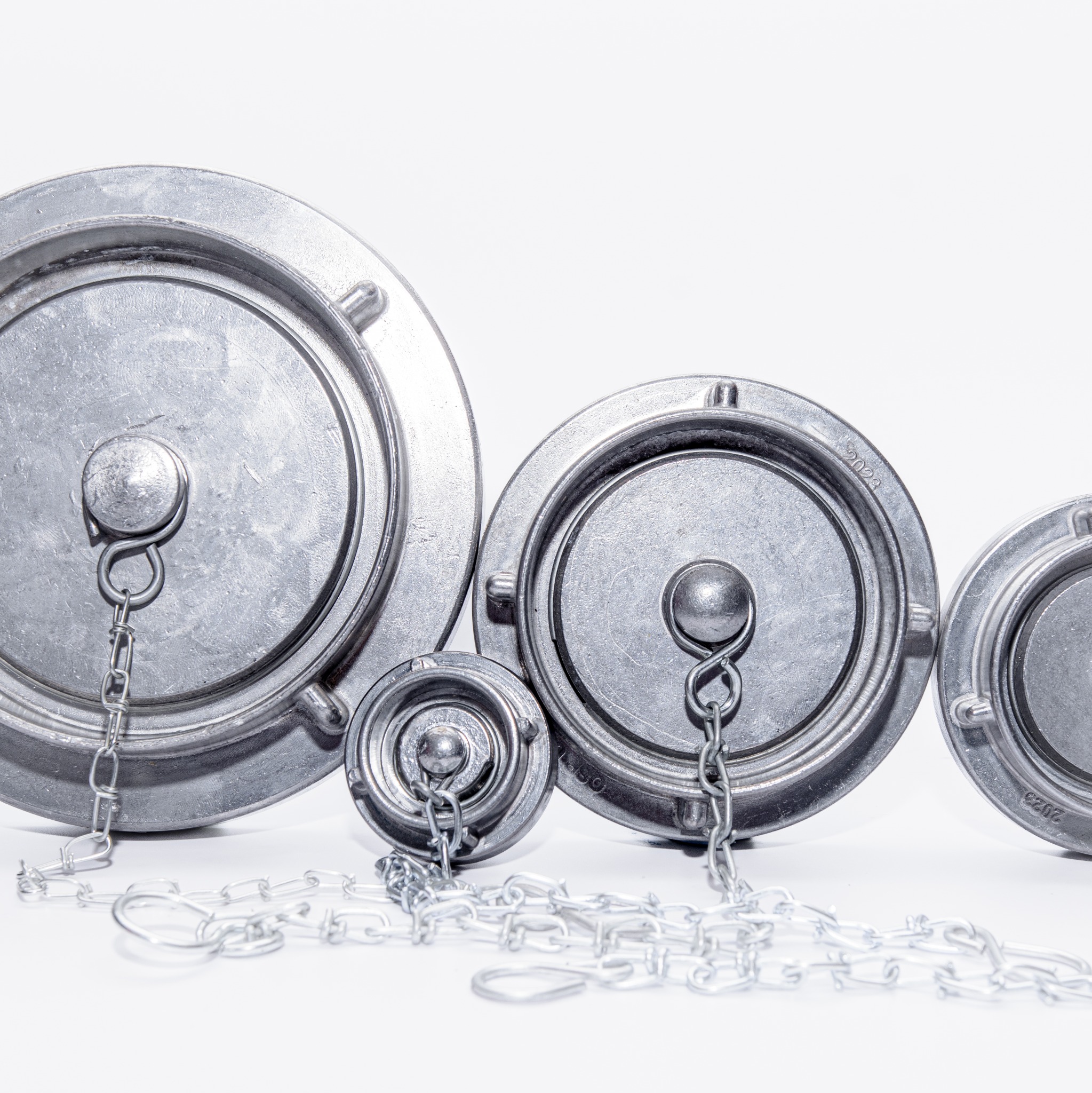 Metalowe, okrągłe pokrywy z łańcuszkami różnych rozmiarów, ułożone obok siebie na białym tle. Widoczne detale wykonania i oznaczenia na powierzchni.