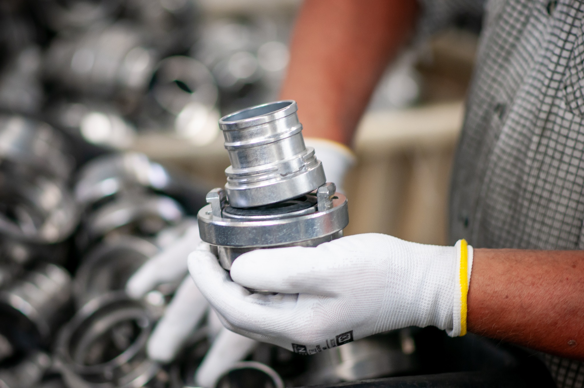 Metalowy łącznik w dłoniach w białych rękawicach na tle stosu podobnych elementów. Detal obróbki metalu, precyzyjne wykonanie.