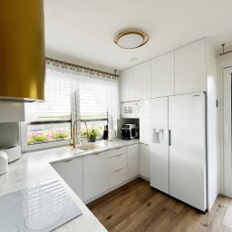 SOKOŁOWSKA architekt wnętrz - Jasna kuchnia z białymi szafkami, marmurowym blatem i złotymi akcentami. Widok na okno z kwiatami i drewnianą podłogę. Nowoczesny design z dbałością o detale.