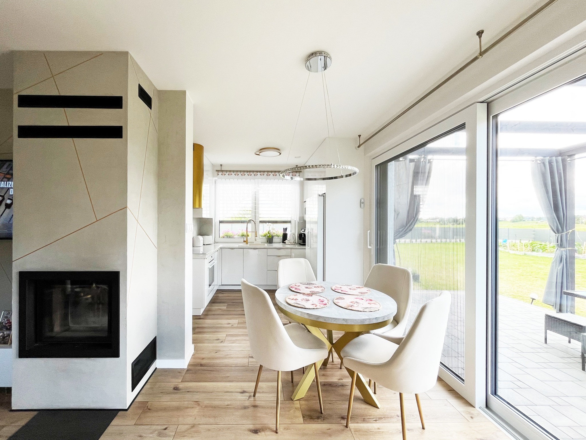 Jasne wnętrze z kominkiem i okrągłym stołem. Widok na kuchnię i taras. Parkiet na podłodze. Nowoczesny design z elementami złota.