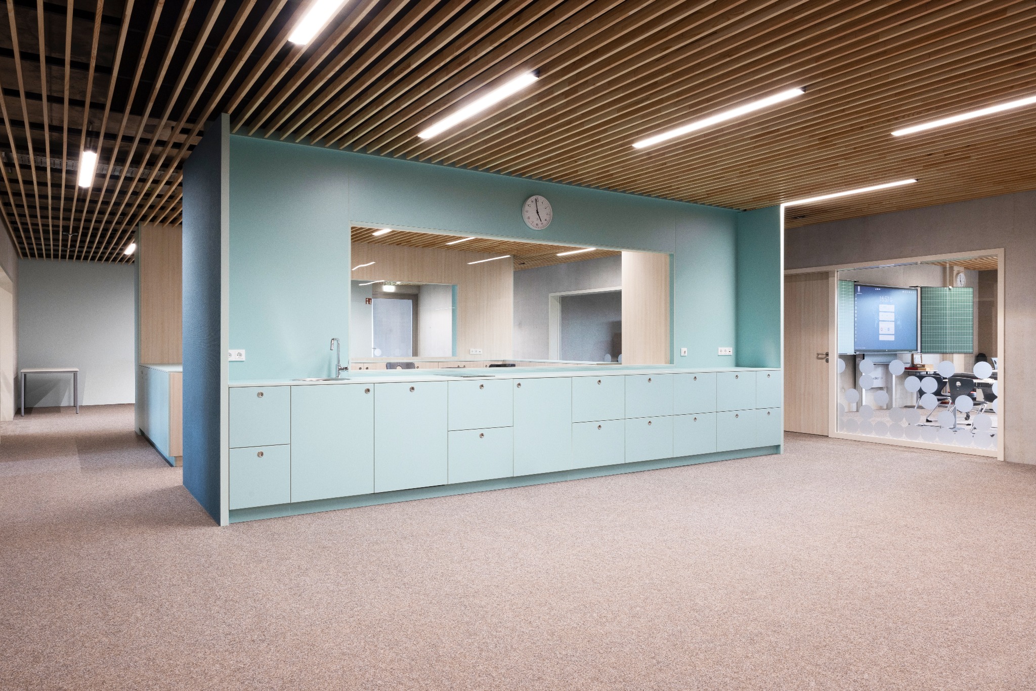 Jasne biuro z zabudową meblową w odcieniach błękitu i beżu, drewnianym sufitem rastrowym i wykładziną dywanową. Widoczny zlew, lustro i zegar.