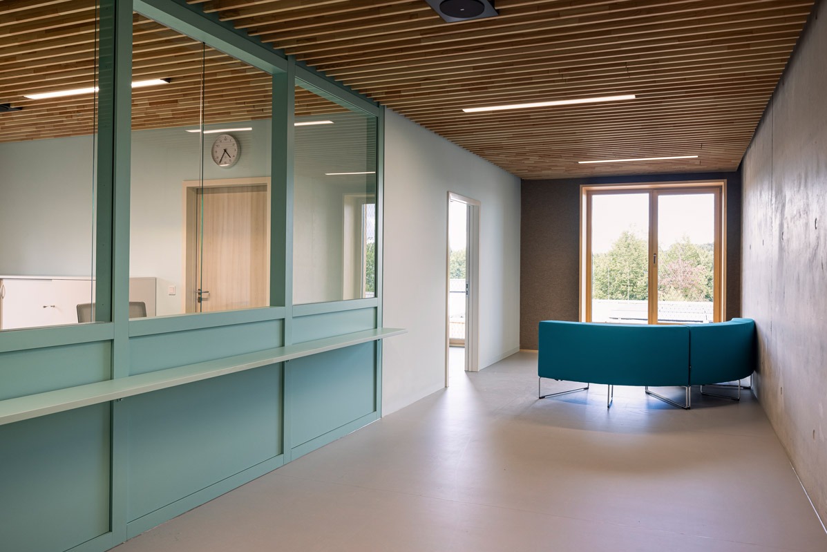 Nowoczesne wnętrze biurowe z turkusową sofą, drewnianym sufitem i przeszkloną recepcją. Minimalistyczny design, jasne kolory, widok na zieleń za oknem.