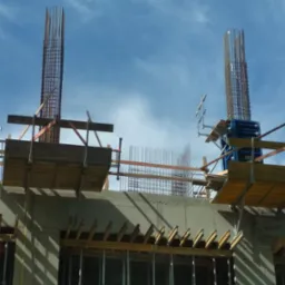 Zbrojenie pionowych słupów na budowie z widocznymi szalunkami i deskowaniem, na tle błękitnego nieba. Etap przygotowawczy do zalewania betonem.