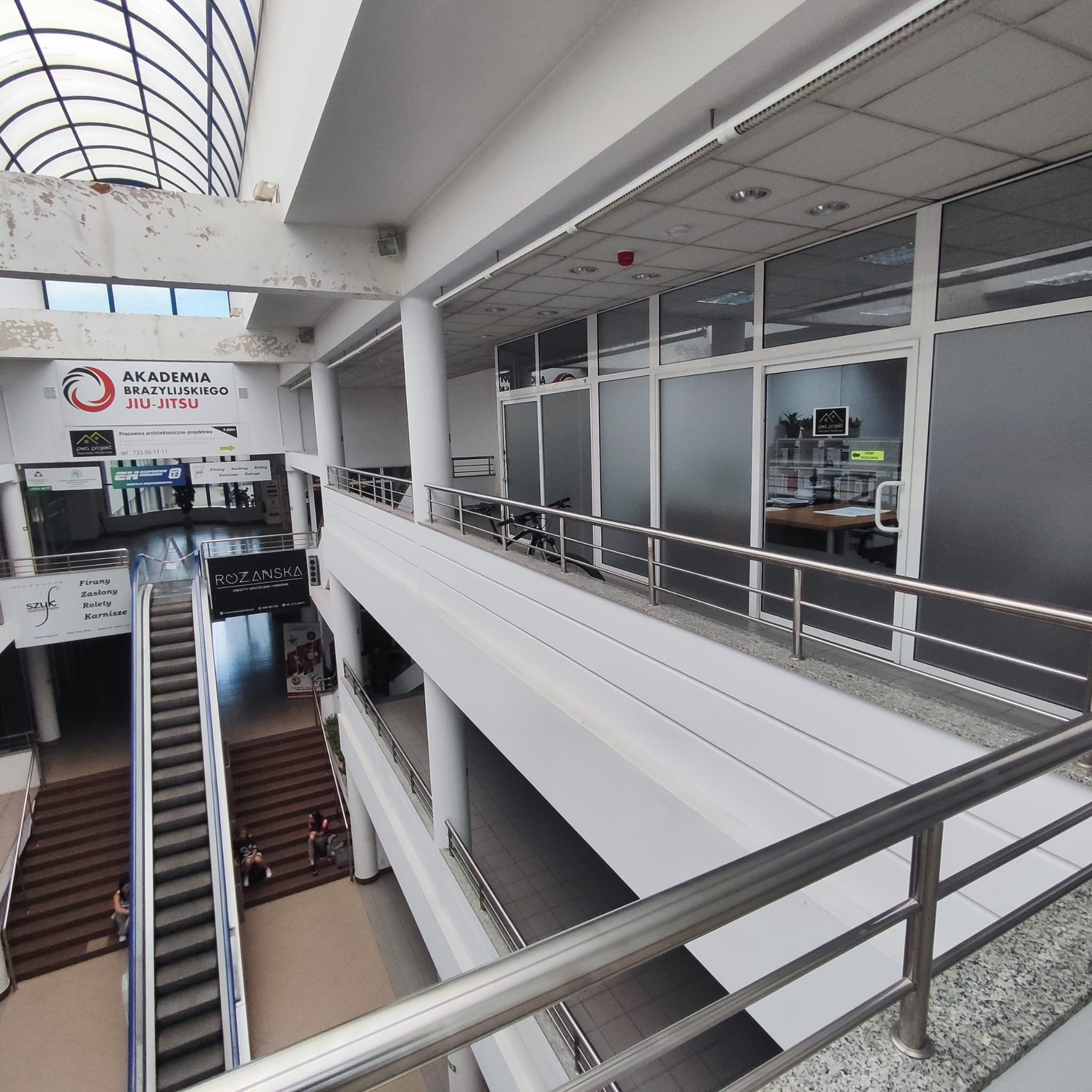 Wnętrze budynku usługowego z widocznymi schodami ruchomymi, balustradami i biurem projektowym z logiem PWA Projekt. Widok z góry na kilka poziomów.