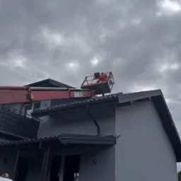 Pracownik w pomarańczowym kombinezonie na podnośniku koszowym, czyszczący dach nowoczesnego, dwupiętrowego budynku z szarej elewacji w pochmurny dzień.