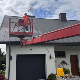 Czysta hata - Pracownik na podnośniku koszowym przy dachu domu z anteną. Widoczny fragment elewacji, brama garażowa i fragment podjazdu z kostki brukowej. Praca na wysokości.