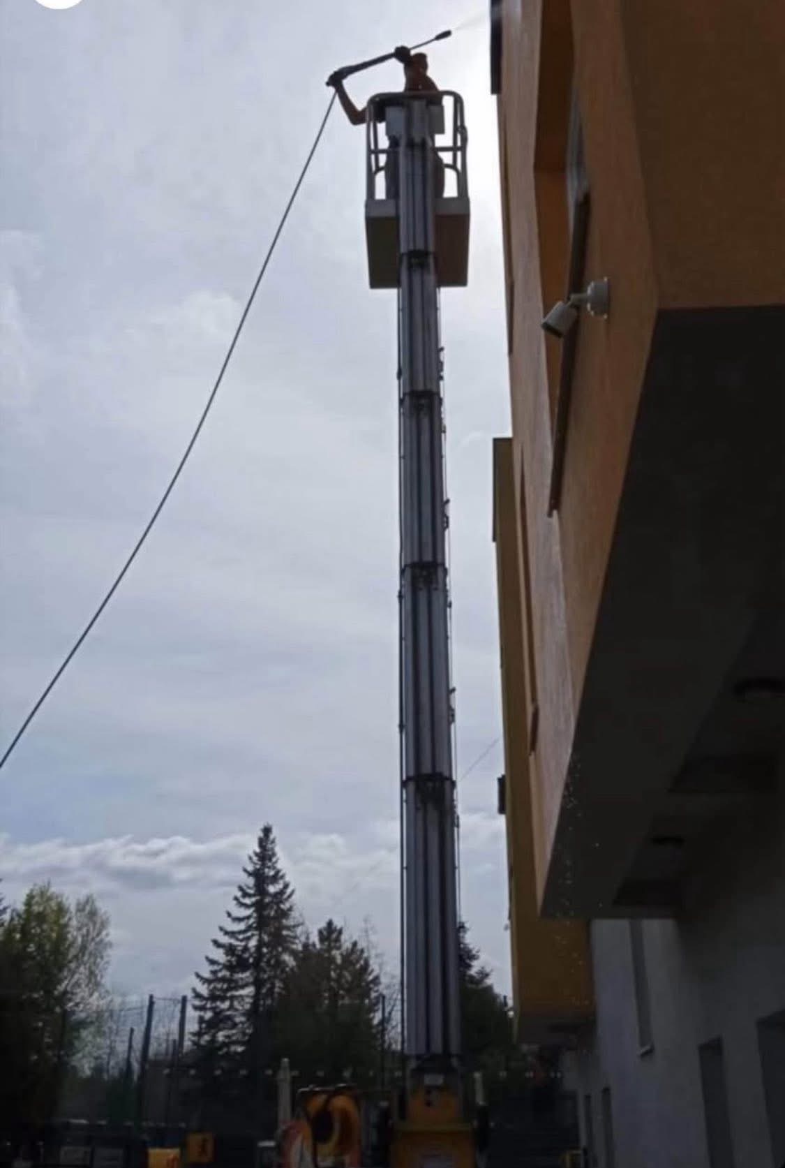Pracownik na podnośniku koszowym, myje elewację budynku z pomarańczową fasadą, używając myjki ciśnieniowej. W tle widoczne drzewa i pochmurne niebo.