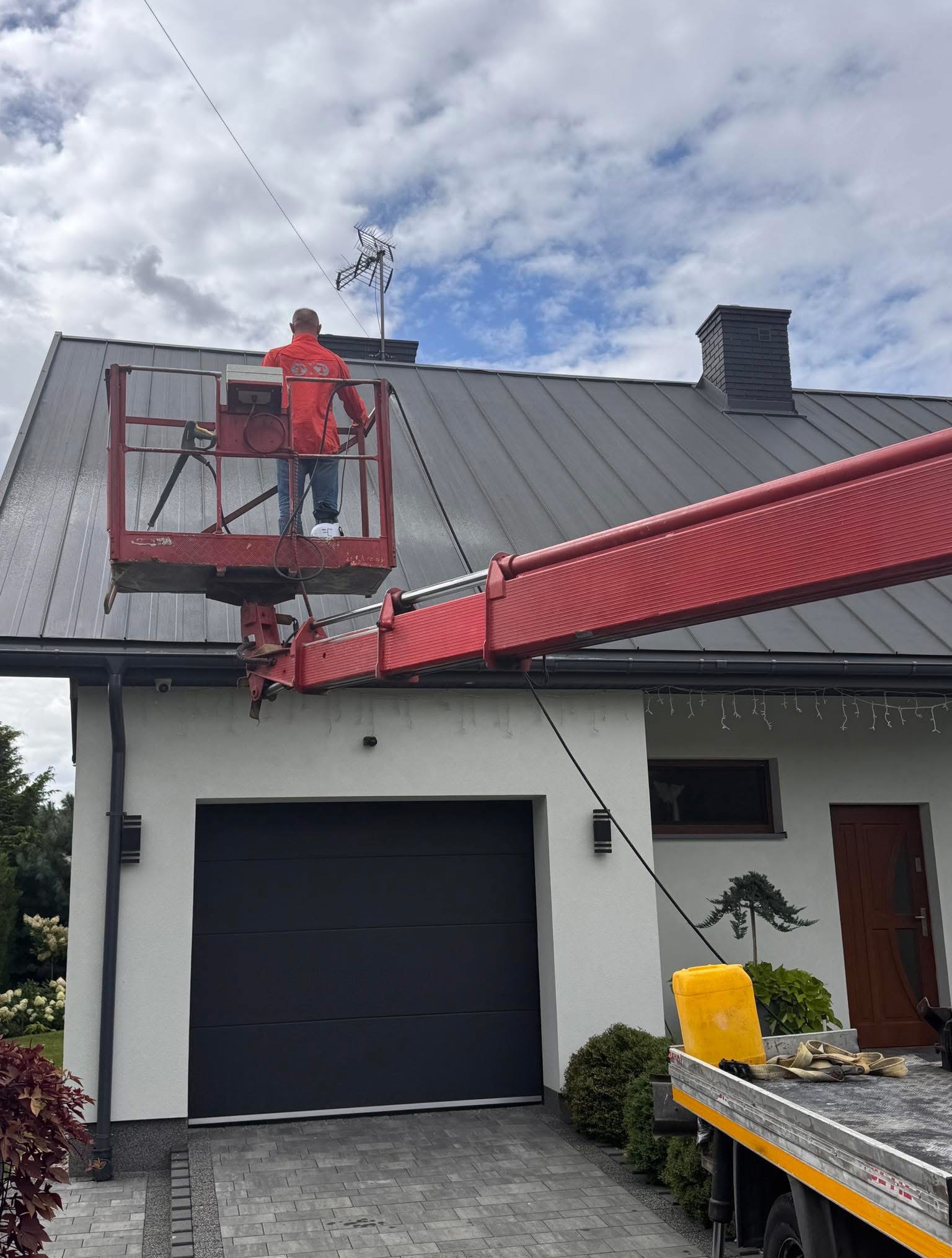 Pracownik na podnośniku koszowym przy dachu domu z anteną. Widoczny fragment elewacji, brama garażowa i fragment podjazdu z kostki brukowej. Praca na wysokości.