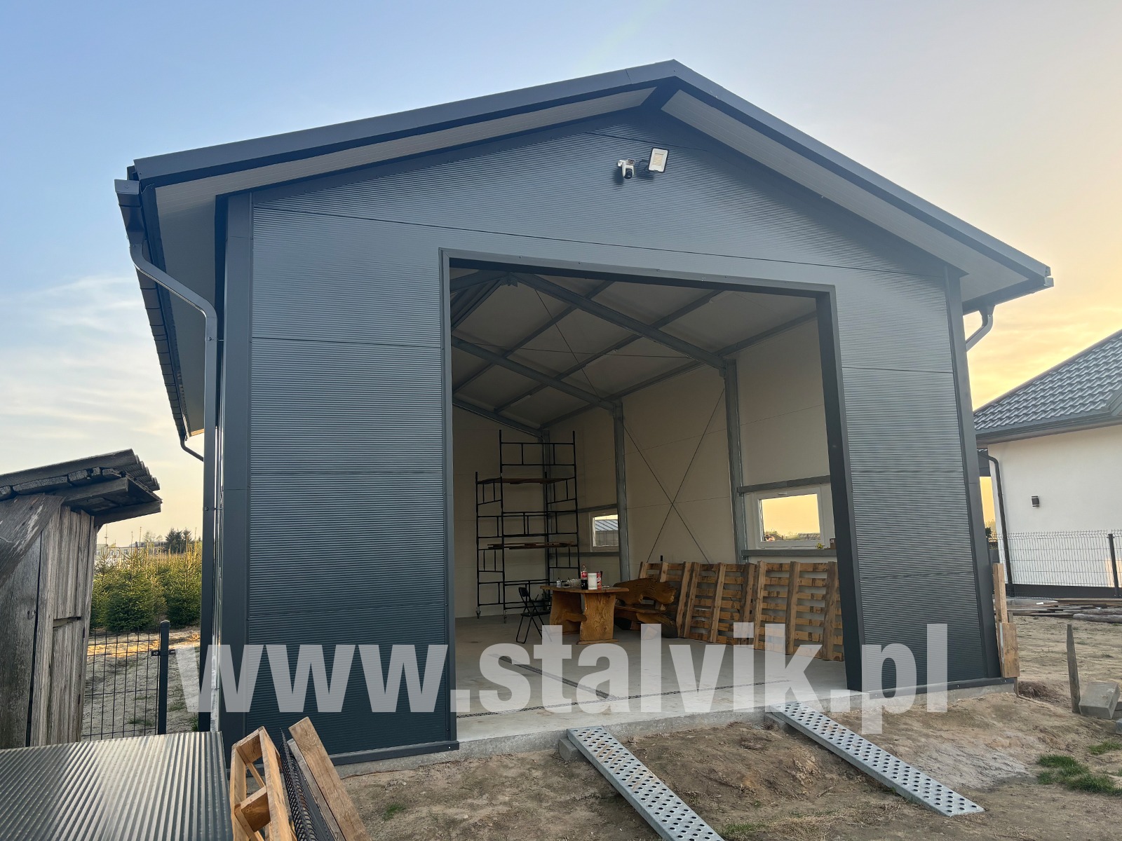 Nowoczesna hala z szarej blachy trapezowej z otwartą bramą wjazdową. Widoczne fragmenty konstrukcji stalowej i okna. Na zewnątrz podjazdy.