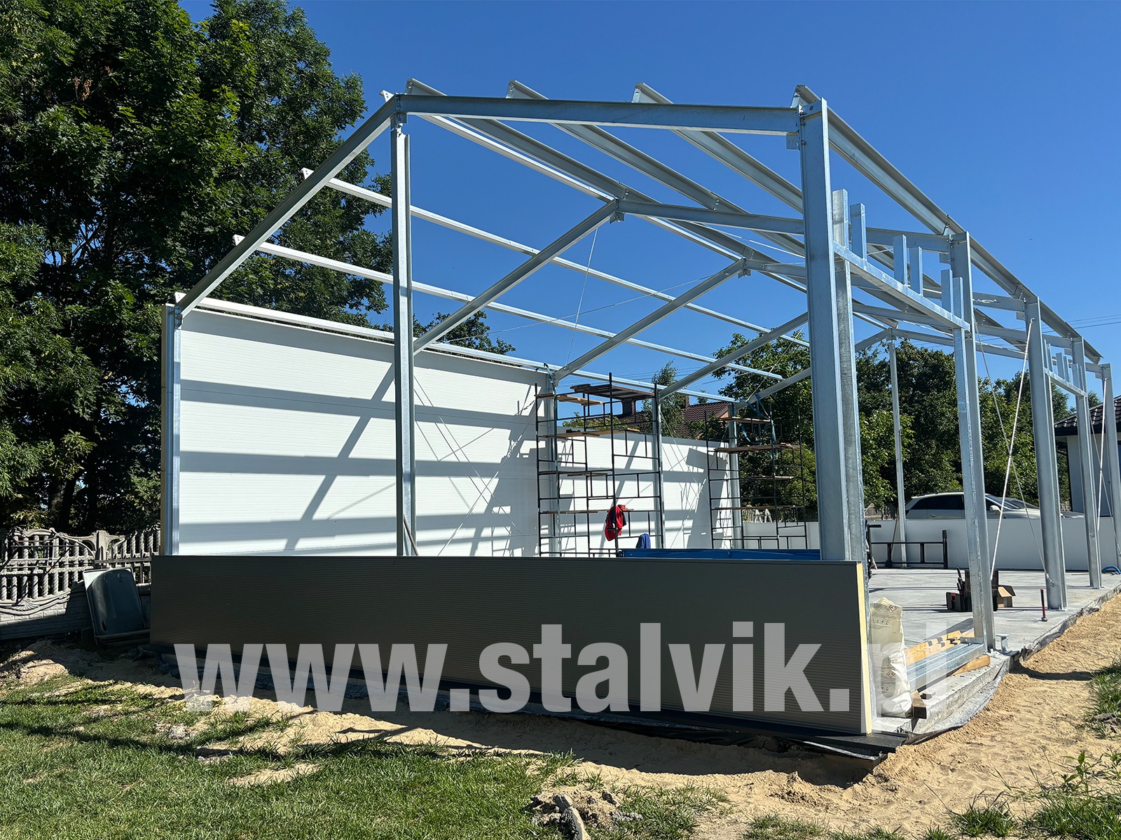 Budowa hali: stalowa konstrukcja szkieletowa z białymi panelami, widoczne rusztowanie. Elementy konstrukcji w surowym, srebrnym kolorze.