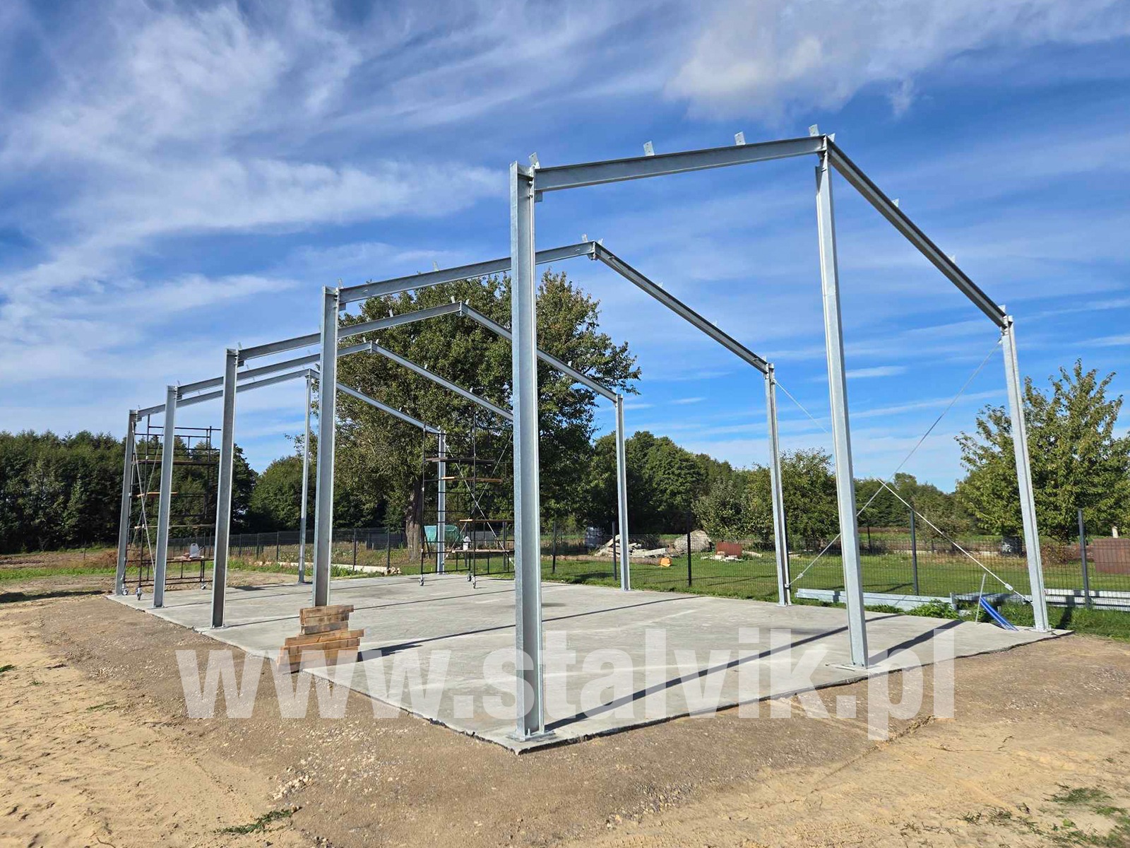 Stalowa konstrukcja hali w trakcie budowy, widoczne rusztowania, zabetonowany fundament, błękitne niebo w tle. Na dole zdjęcia adres strony internetowej.