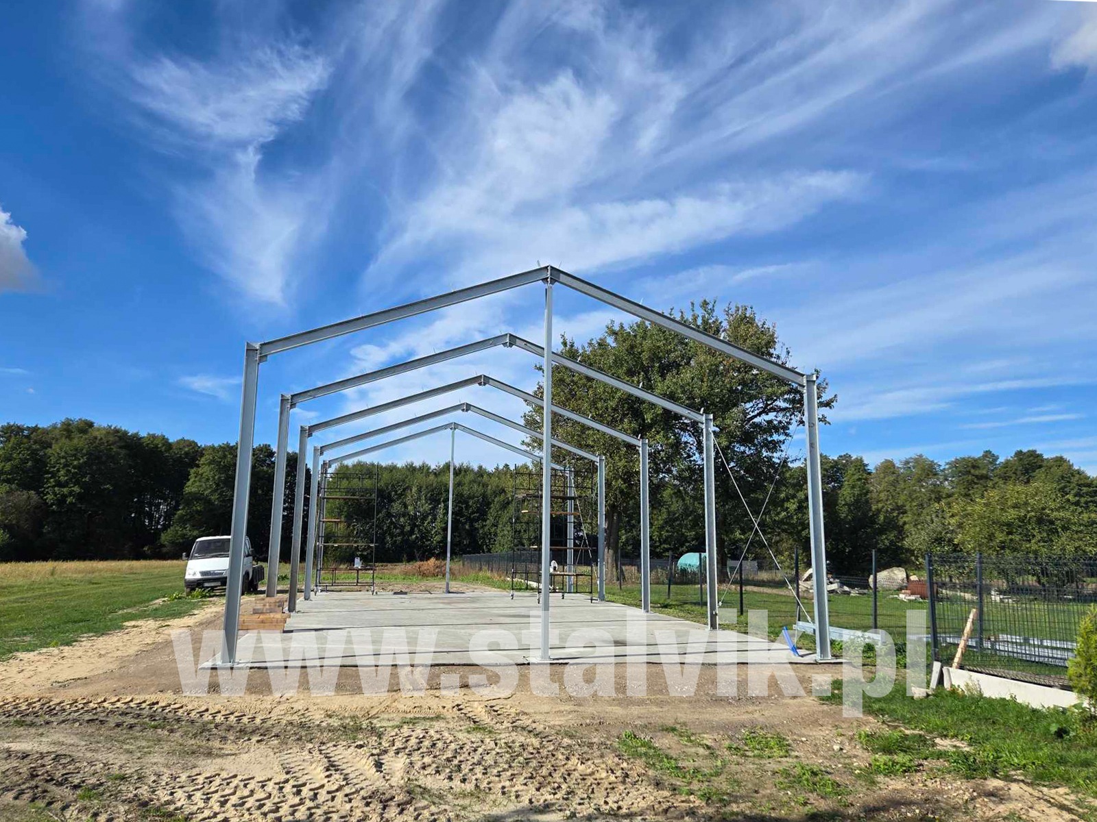 Wznoszenie szkieletu hali stalowej na betonowej płycie fundamentowej, widok z zewnątrz. Konstrukcja na tle błękitnego nieba i zieleni drzew.