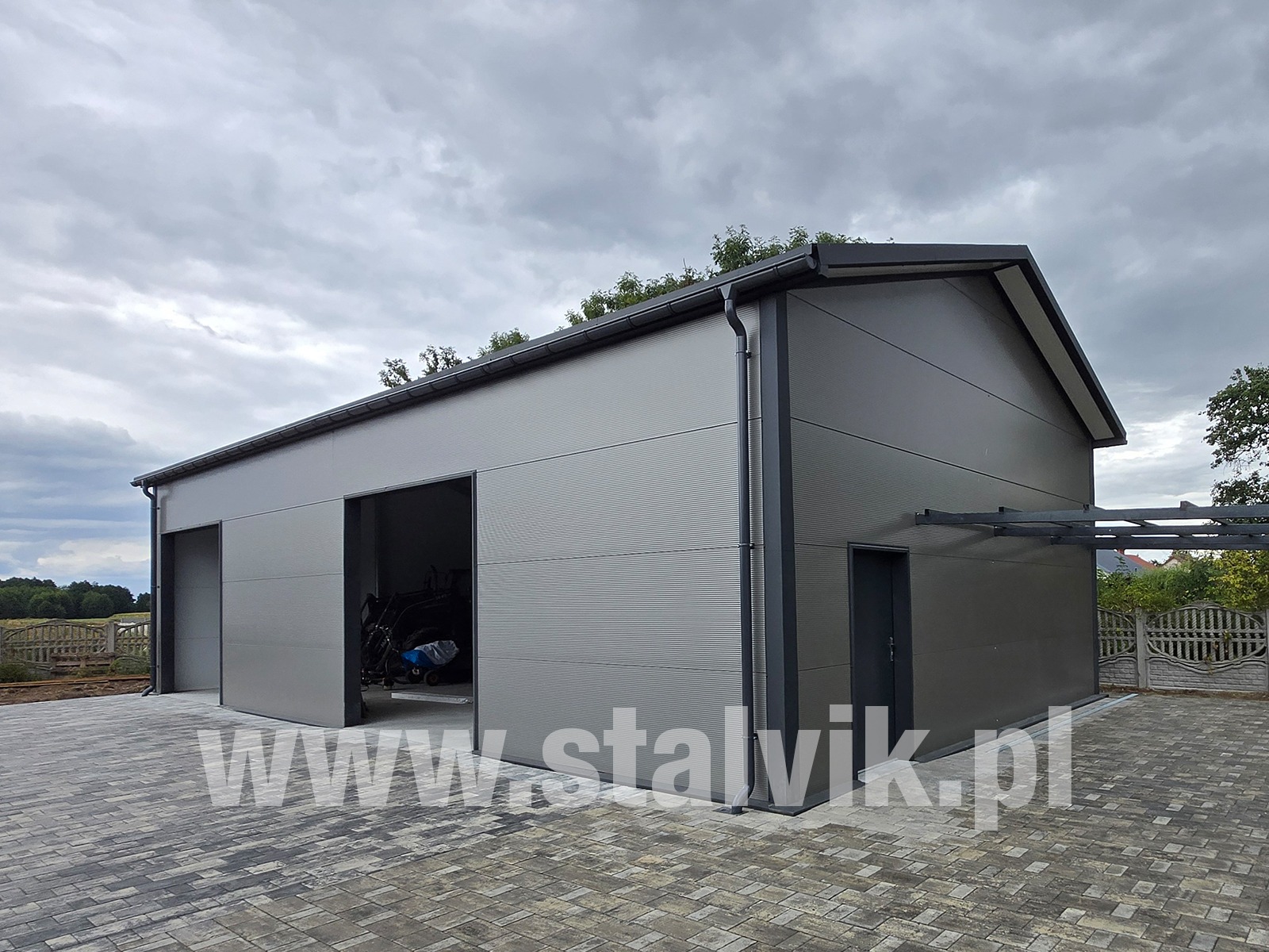 Szara hala z płyty warstwowej z otwartymi wrotami garażowymi, widok z zewnątrz na kostce brukowej. Widoczne detale wykończenia i orynnowanie.