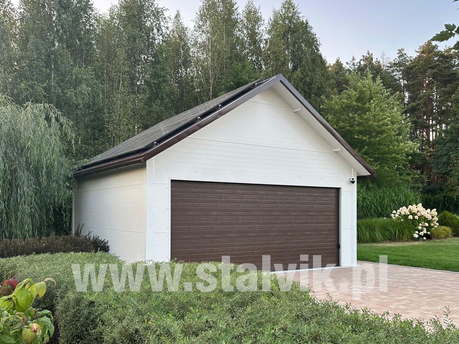 Biała hala z brązową bramą segmentową, otoczona zielenią. Na dachu widoczne panele solarne. Uwagę zwraca estetyczne wykończenie i wkomponowanie w otoczenie.