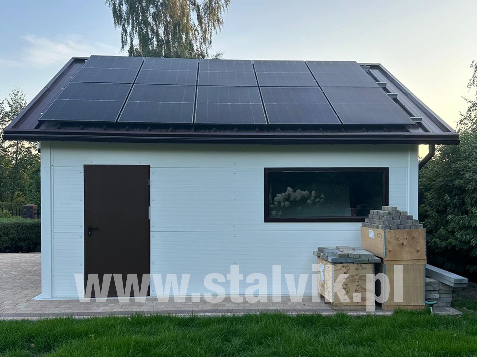 Gotowa hala z paneli warstwowych z panelami solarnymi na dachu. Obok ułożone kostki brukowe. Widoczny adres strony internetowej firmy.
