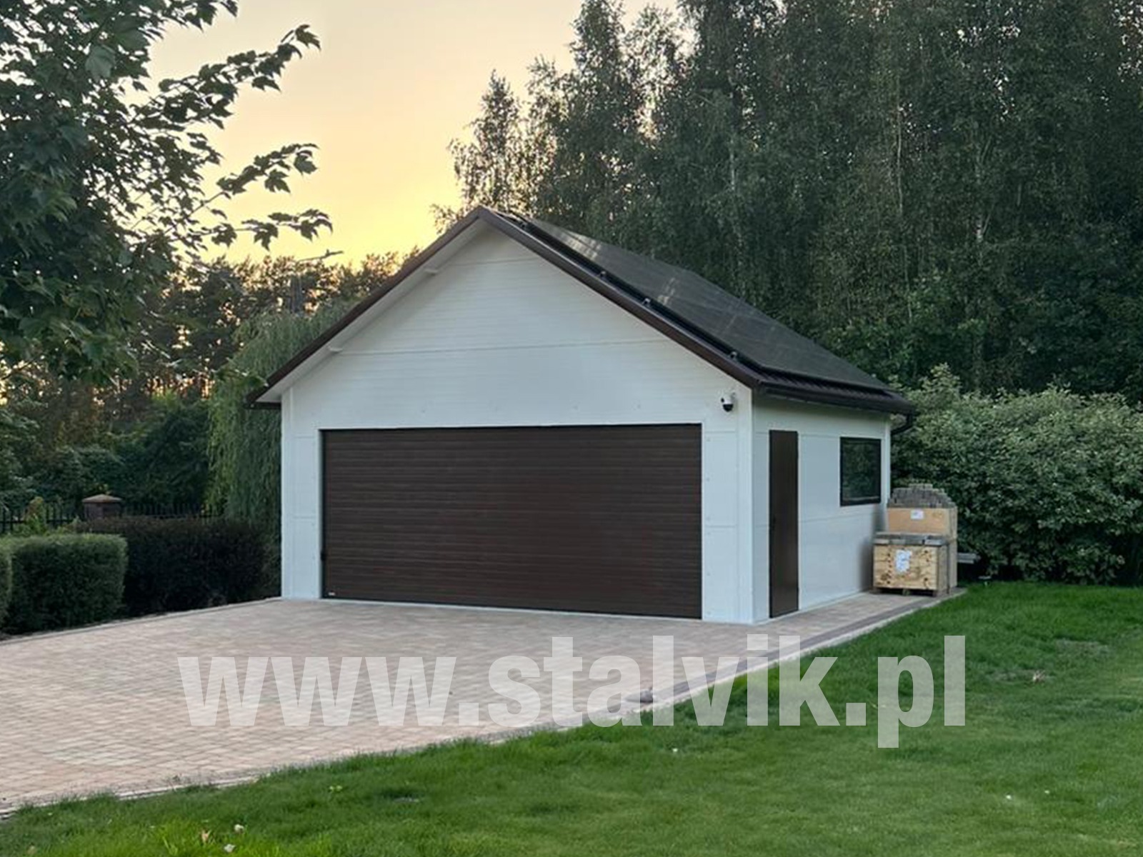 Biała hala z brązową bramą garażową i panelami słonecznymi na dachu, otoczona zielenią. Widok na elewację i podjazd z kostki.