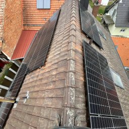 APOLLO SUN SPÓŁKA Z OGRANICZONĄ ODPOWIEDZIALNOŚCIĄ - Montaż paneli fotowoltaicznych na dachu z ceramiczną dachówką. Widok z góry, panele ułożone w rzędach, częściowo widoczne buty montera na dole kadru.