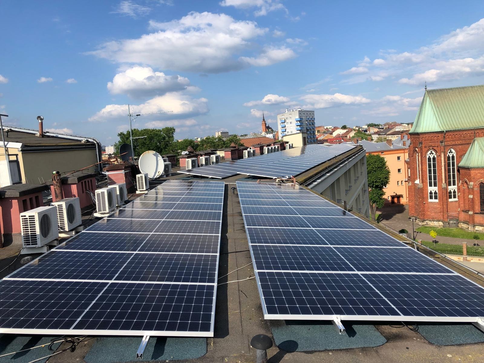 Instalacja paneli fotowoltaicznych na dachu budynku z klimatyzatorami i anteną satelitarną, w tle zabudowa miejska i błękitne niebo z chmurami.