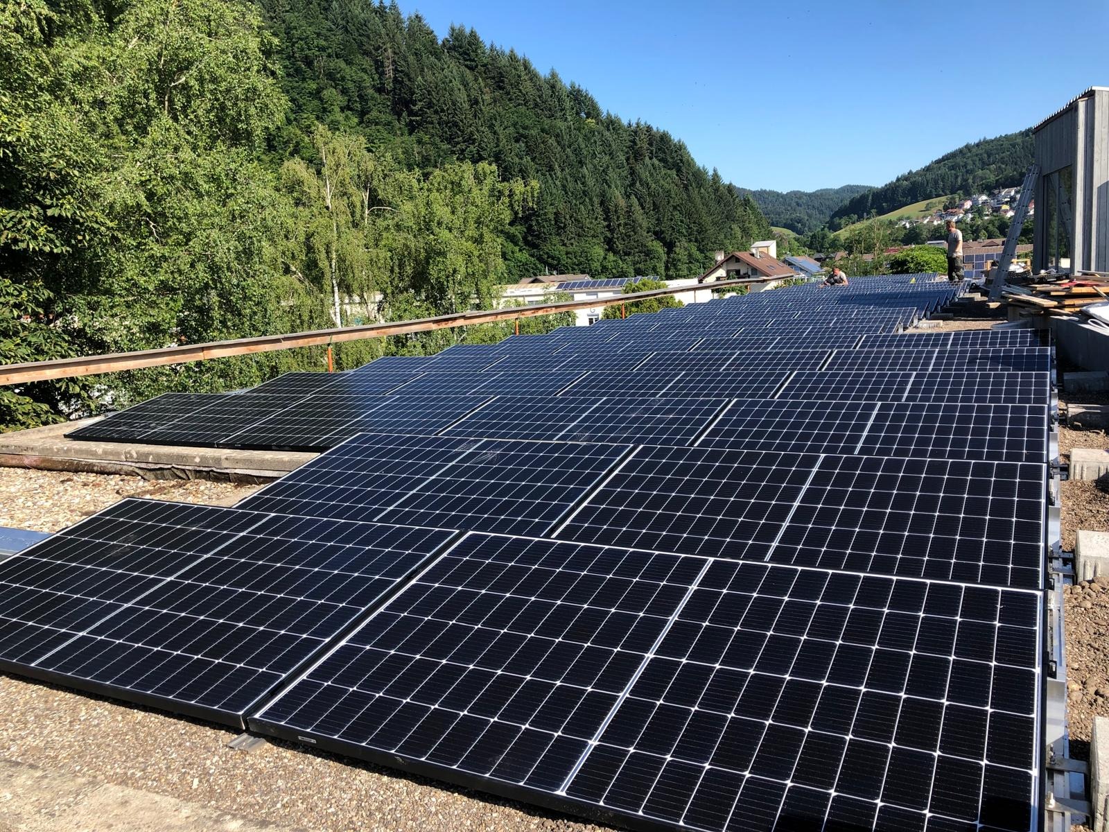 Instalacja paneli fotowoltaicznych na dachu budynku, z widokiem na zielone wzgórza w tle. Widoczni pracownicy podczas montażu.