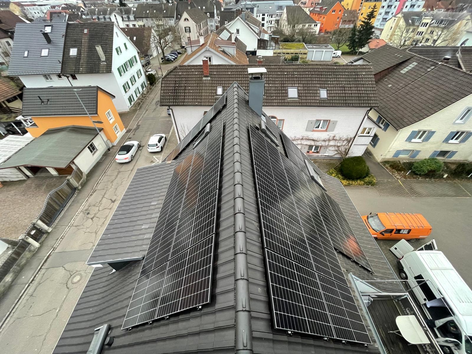 Widok z góry na dach z panelami słonecznymi na tle zabudowy miejskiej. Pomarańczowy samochód dostawczy zaparkowany obok białego. Dwa białe samochody na ulicy.