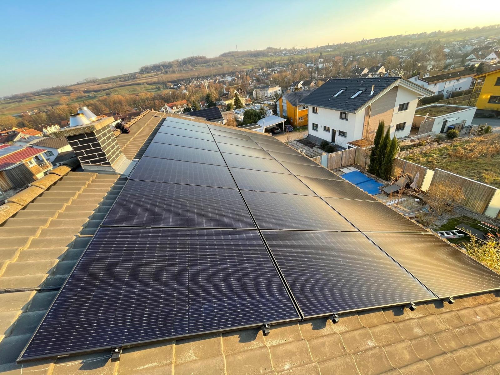 Panele fotowoltaiczne na dachu domu jednorodzinnego, widok z góry, w tle zabudowa mieszkalna i pagórkowaty krajobraz w słoneczny dzień.