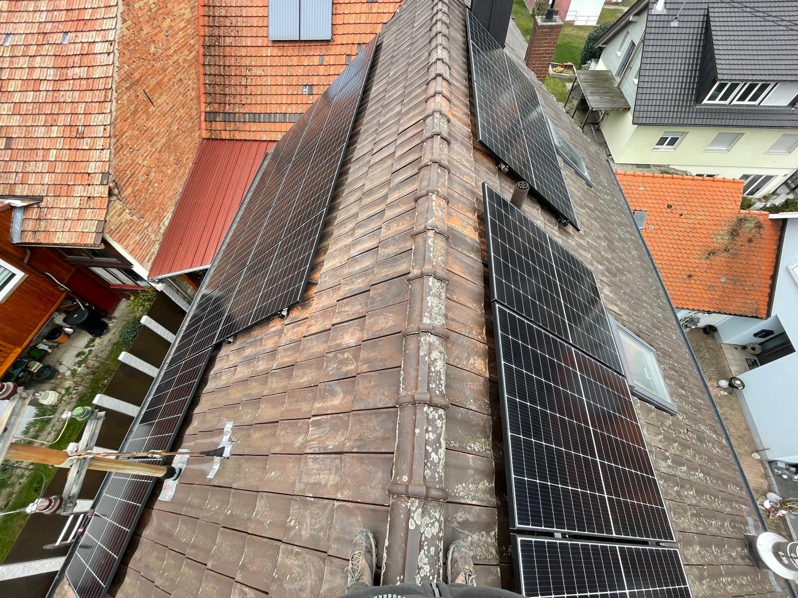 Montaż paneli fotowoltaicznych na dachu z ceramiczną dachówką. Widok z góry, panele ułożone w rzędach, częściowo widoczne buty montera na dole kadru.