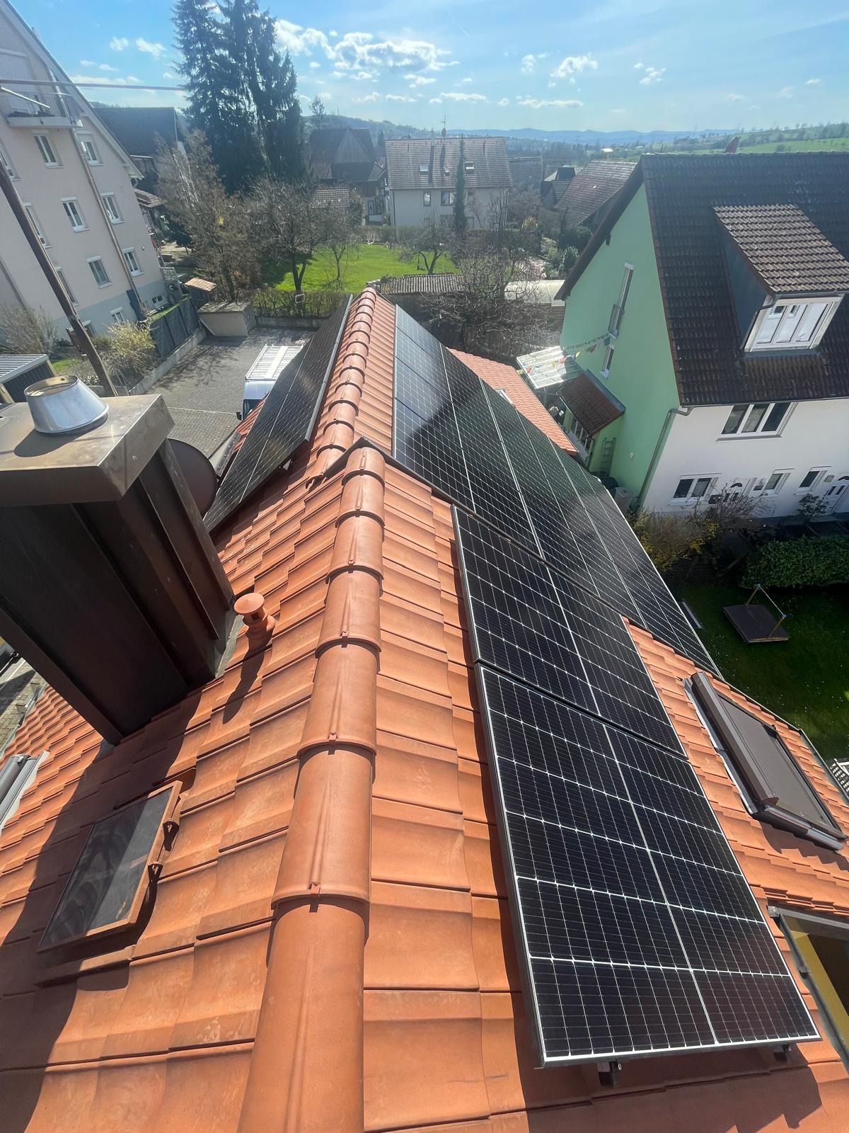 Instalacja paneli słonecznych na dachu z dachówki ceramicznej, widok z góry. W tle zabudowa jednorodzinna i zieleń. Widoczne kominy i okna dachowe.